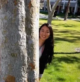 A young woman with long black hair peeking from behind a tree in a park with green grass and trees in the background.