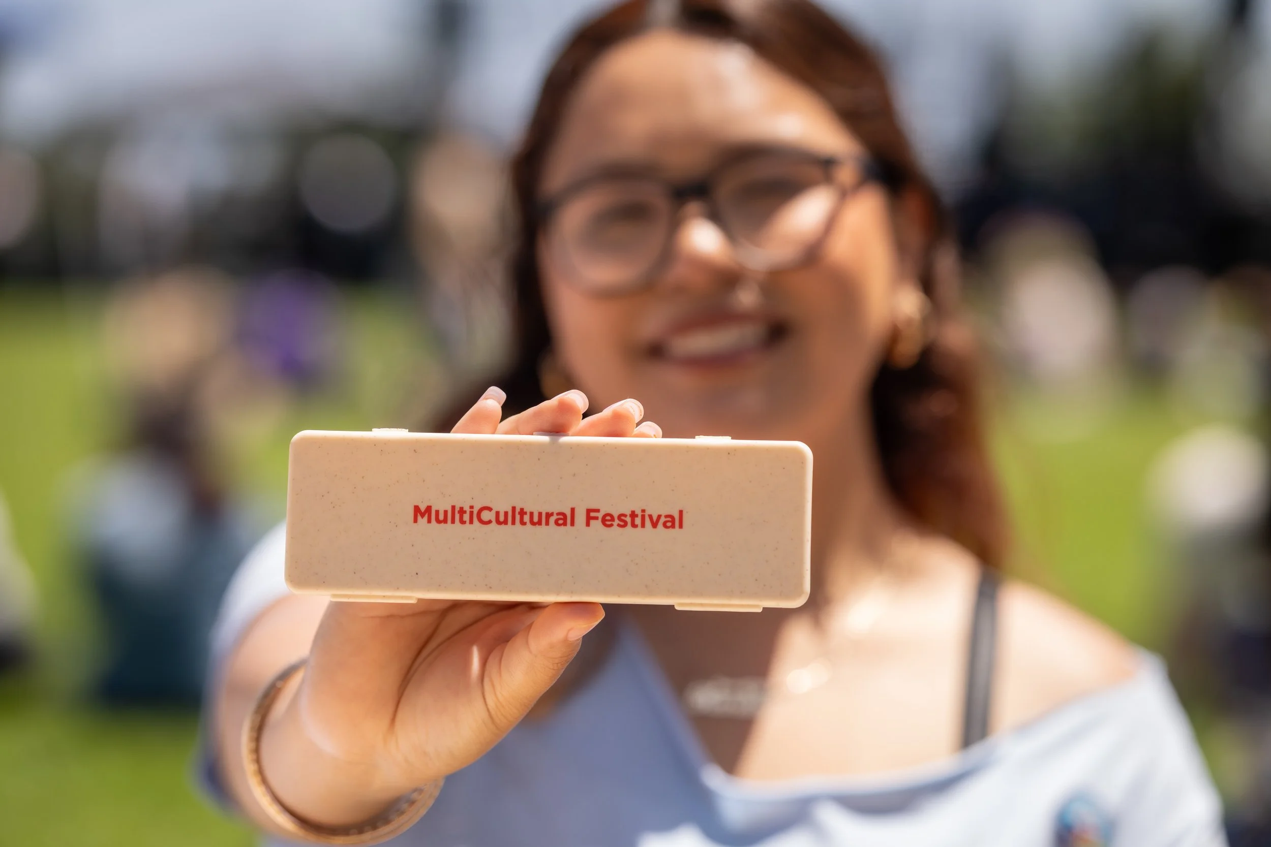 A woman with glasses holding a sign that reads 'MultiCultural Festival' outside in a park or outdoor area on a sunny day.