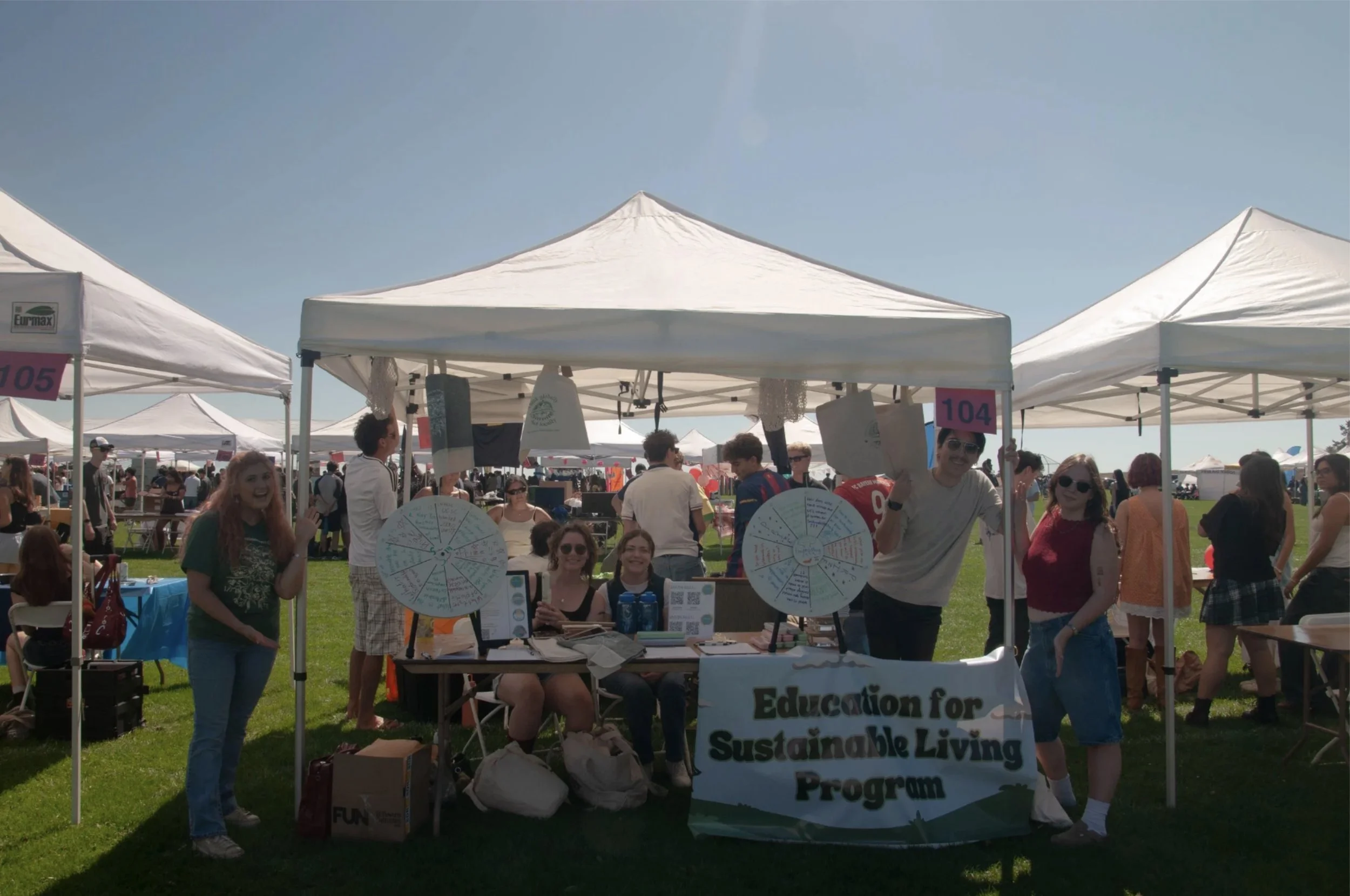 A group of people at an outdoor event, standing under white tents with booths and signs, including one that reads "Education for Sustainable Living Program." The crowd appears cheerful under a clear blue sky.