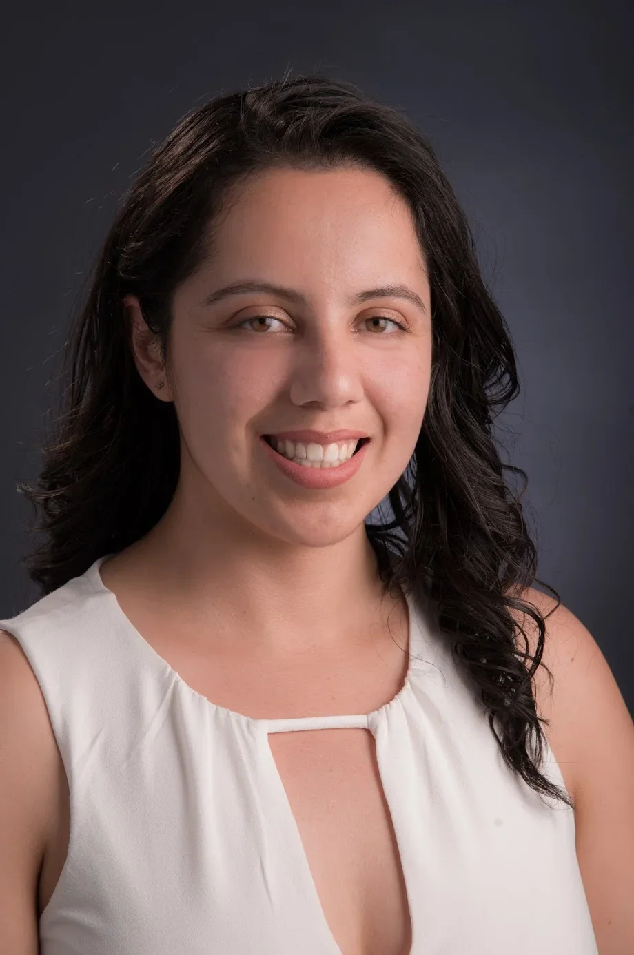 Headshot of a woman with dark wavy hair, smiling, wearing a white sleeveless top, against a dark background.