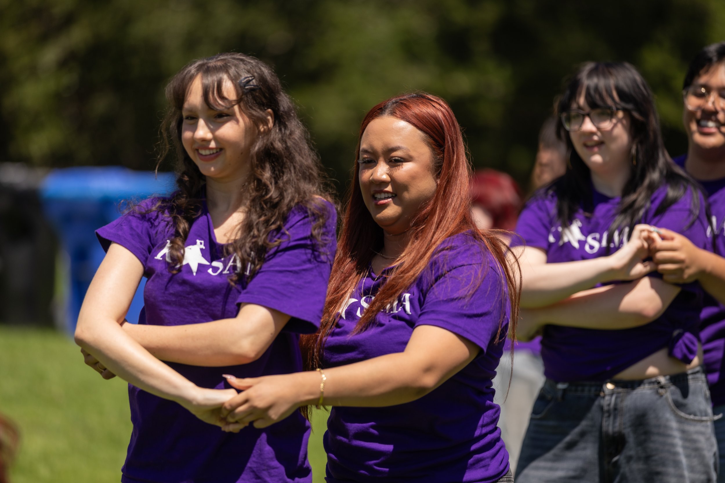 Group of women standing outdoors in a line, wearing purple shirts, smiling, and with arms crossed on a sunny day.