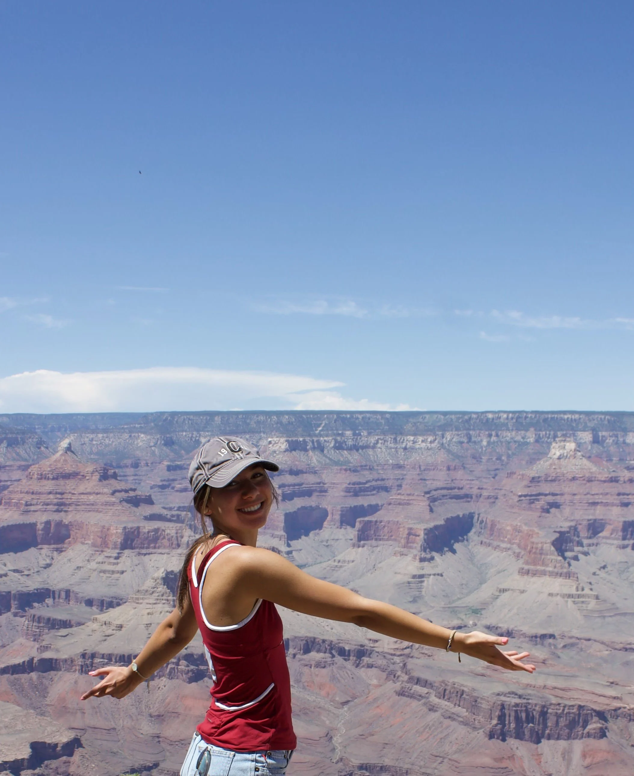 Young woman smiling at the Grand Canyon with arms outstretched, wearing a gray cap, red tank top, and denim shorts.