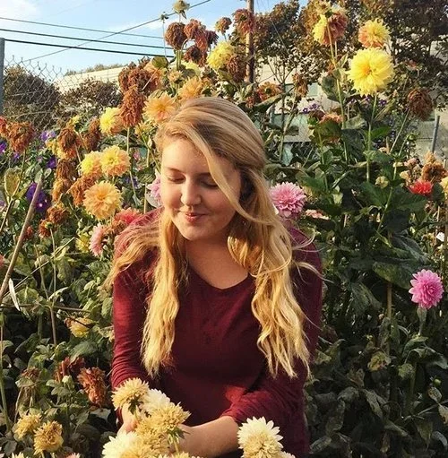 A young woman with long blonde hair, wearing a maroon sweater, sitting among colorful flowers in a garden.