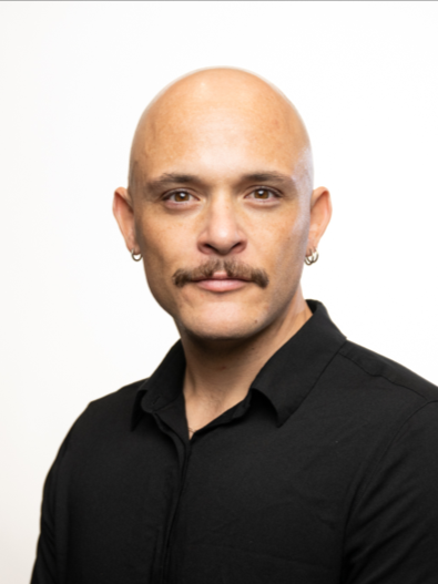 A man with a bald head, mustache, and earrings wearing a black shirt, posing against a plain white background.
