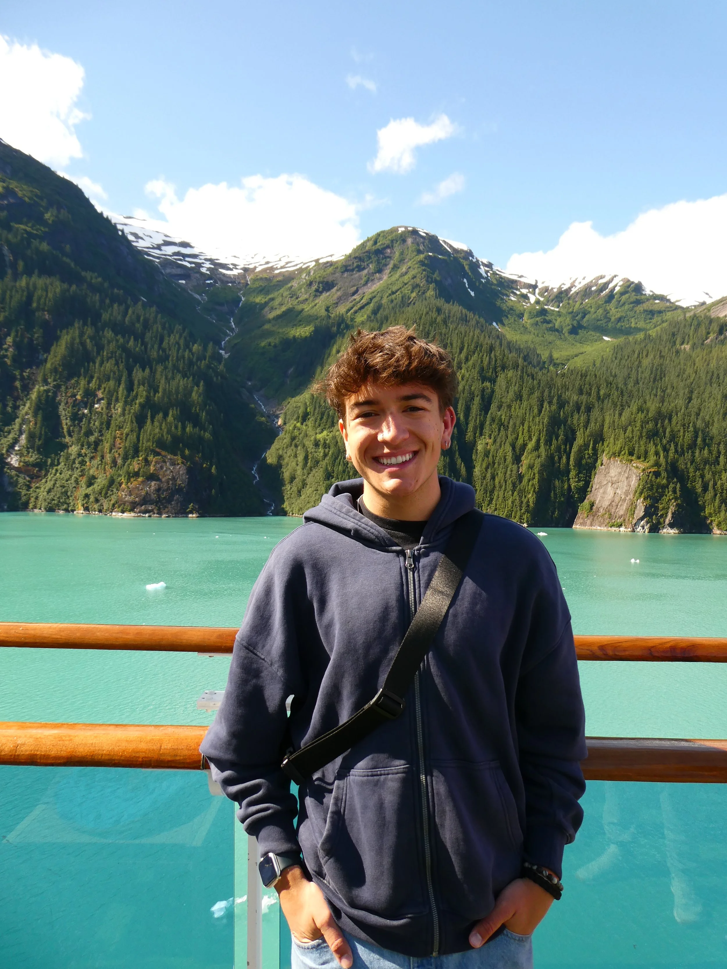 A young man sitting on rocks outdoors with mountains in the background, smiling, wearing a brown shirt with ginkgo leaves and blue jeans.