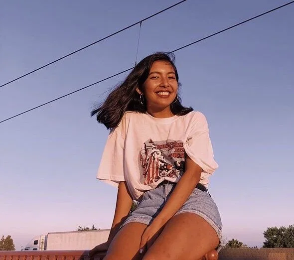 A young woman with long black hair smiling outdoors against a clear evening sky, sitting on a surface with power lines above.
