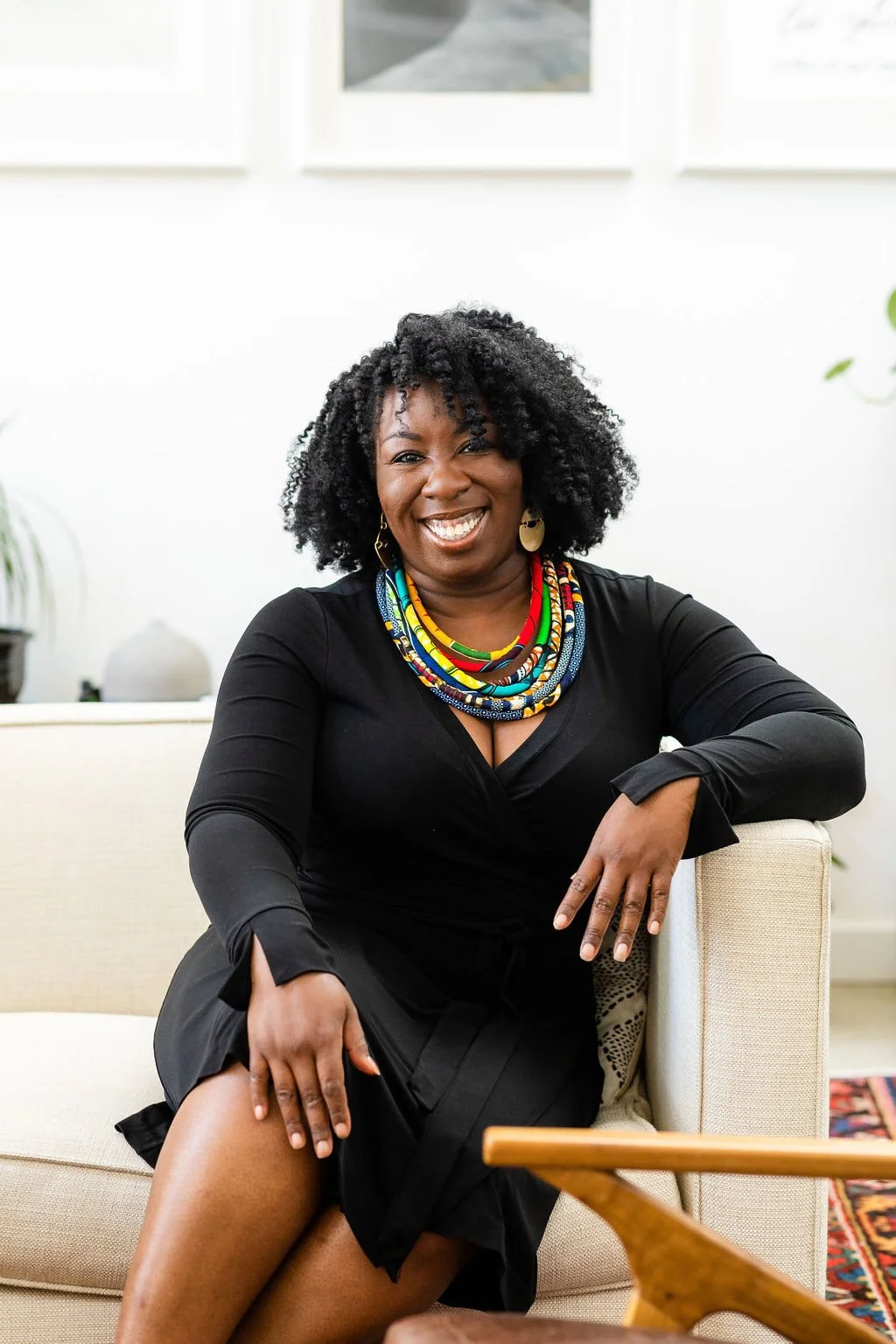 Woman with curly hair smiling, wearing a black dress and colorful beaded necklace, sitting on a beige sofa in a bright room with framed art on the wall.