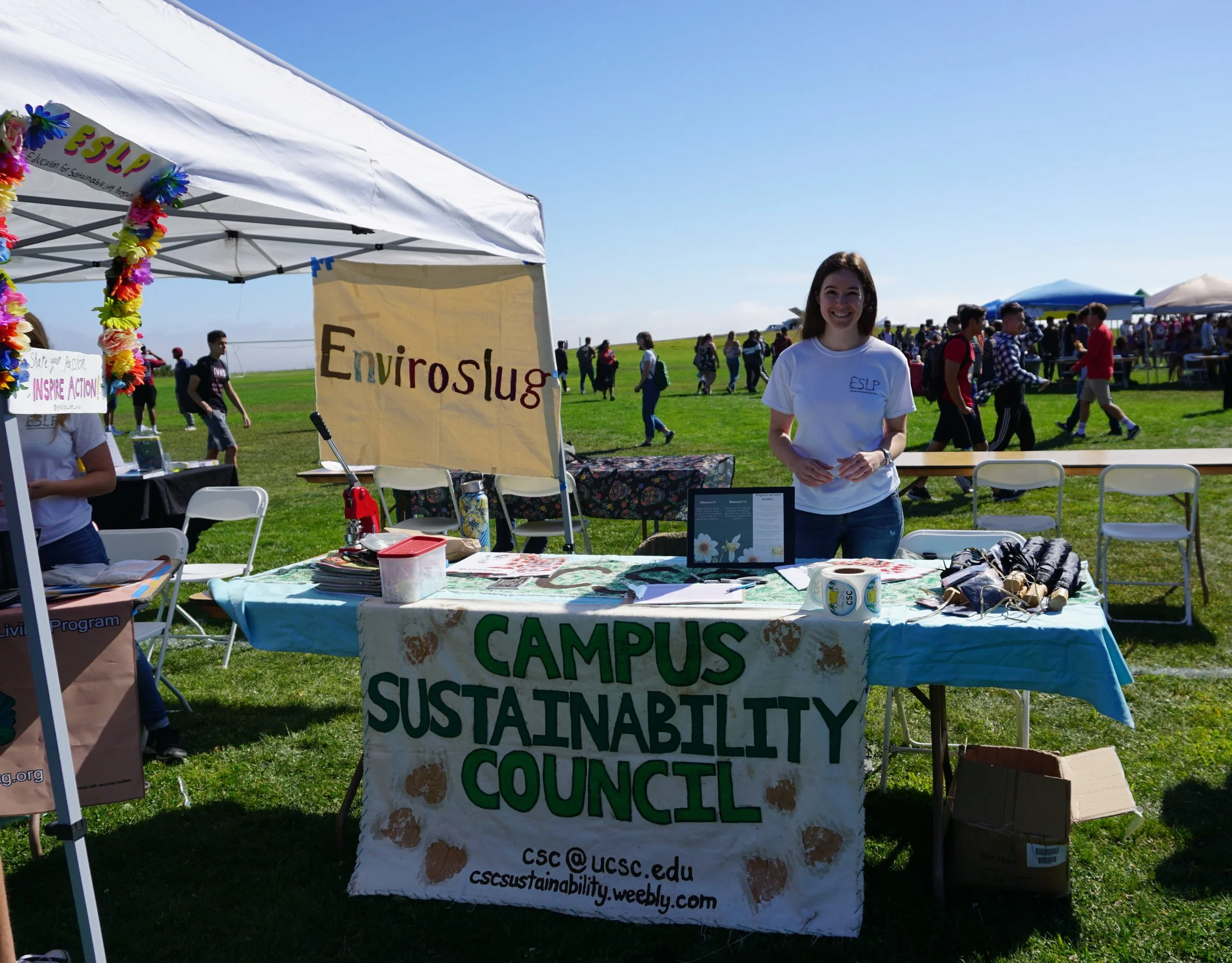 A young woman standing at a table with posters and materials promoting sustainability. Behind her is a sign reading 'EnviroSlog' and another sign with partially visible text about sustainability council. The scene is outdoors on a sunny day with many people and tents in the background.
