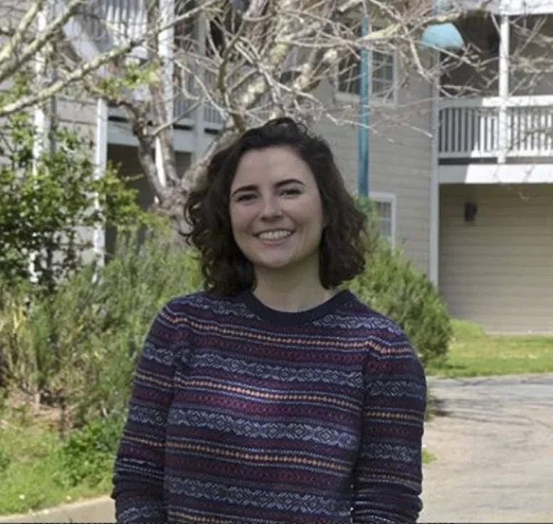 Smiling woman with shoulder-length curly hair standing outdoors in front of a leafless tree and a residential building with balconies, wearing a colorful patterned sweater.