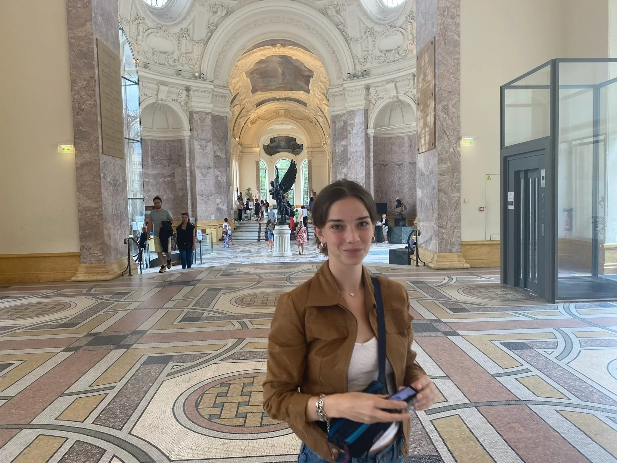 A young woman with short hair and earrings standing inside a grand museum hall with ornate white and pink marble walls, large windows, sculptures, and statues in the background. She is holding a phone and wearing a brown jacket.