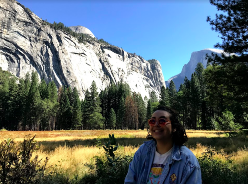 A woman smiling in a denim jacket standing in a grassy field with tall trees and majestic granite cliffs and mountains in the background under a blue sky.