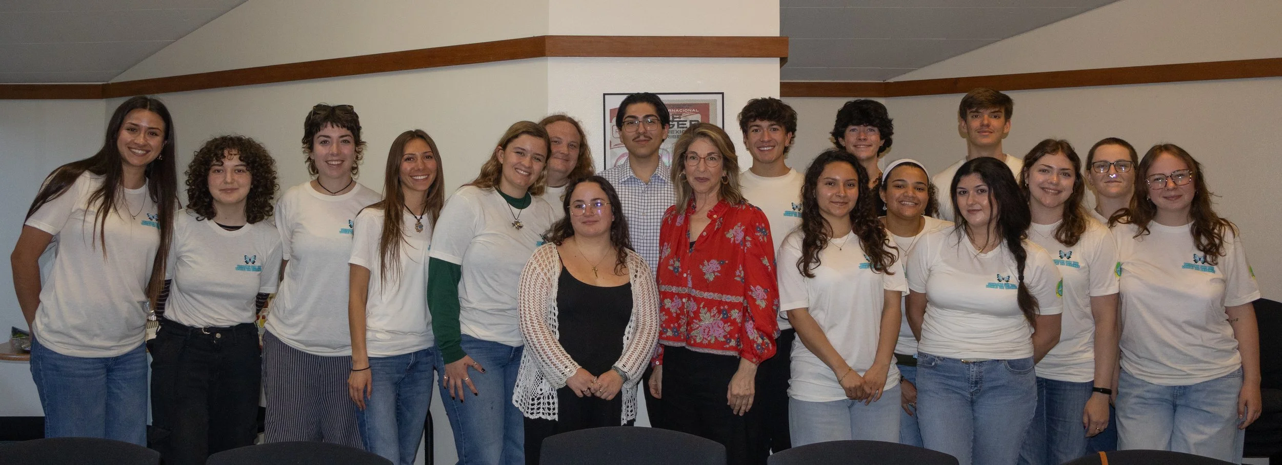 Group of young people and adults posing for a photo indoors, some wearing white T-shirts with blue logos, standing in front of a white wall with framed picture.