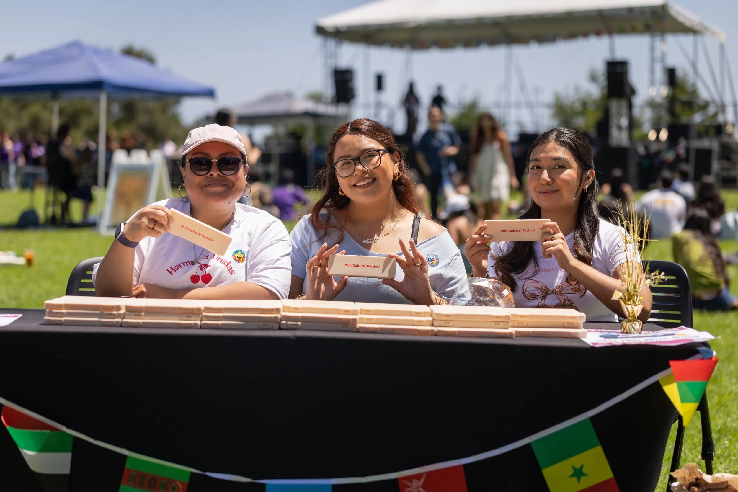 Three women smiling and holding small packages labeled 'Multicultural Festival,' sitting behind a table with a stack of white items. The women are outdoors at a festival with a stage, other people, and a tent visible in the background.