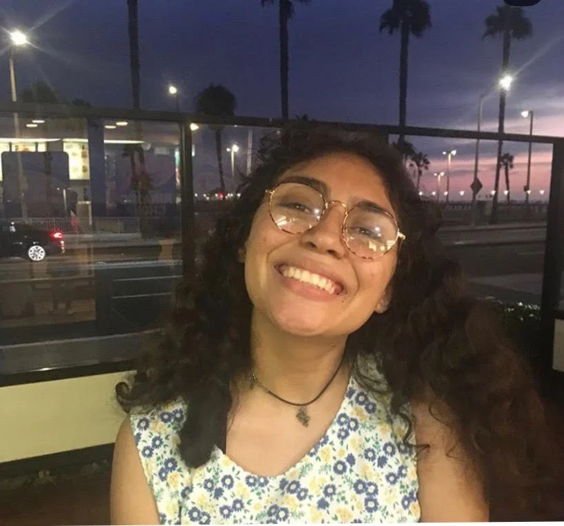 Young woman with curly hair and glasses smiling at the camera inside a restaurant or cafe, with large windows and a sunset sky visible outside.