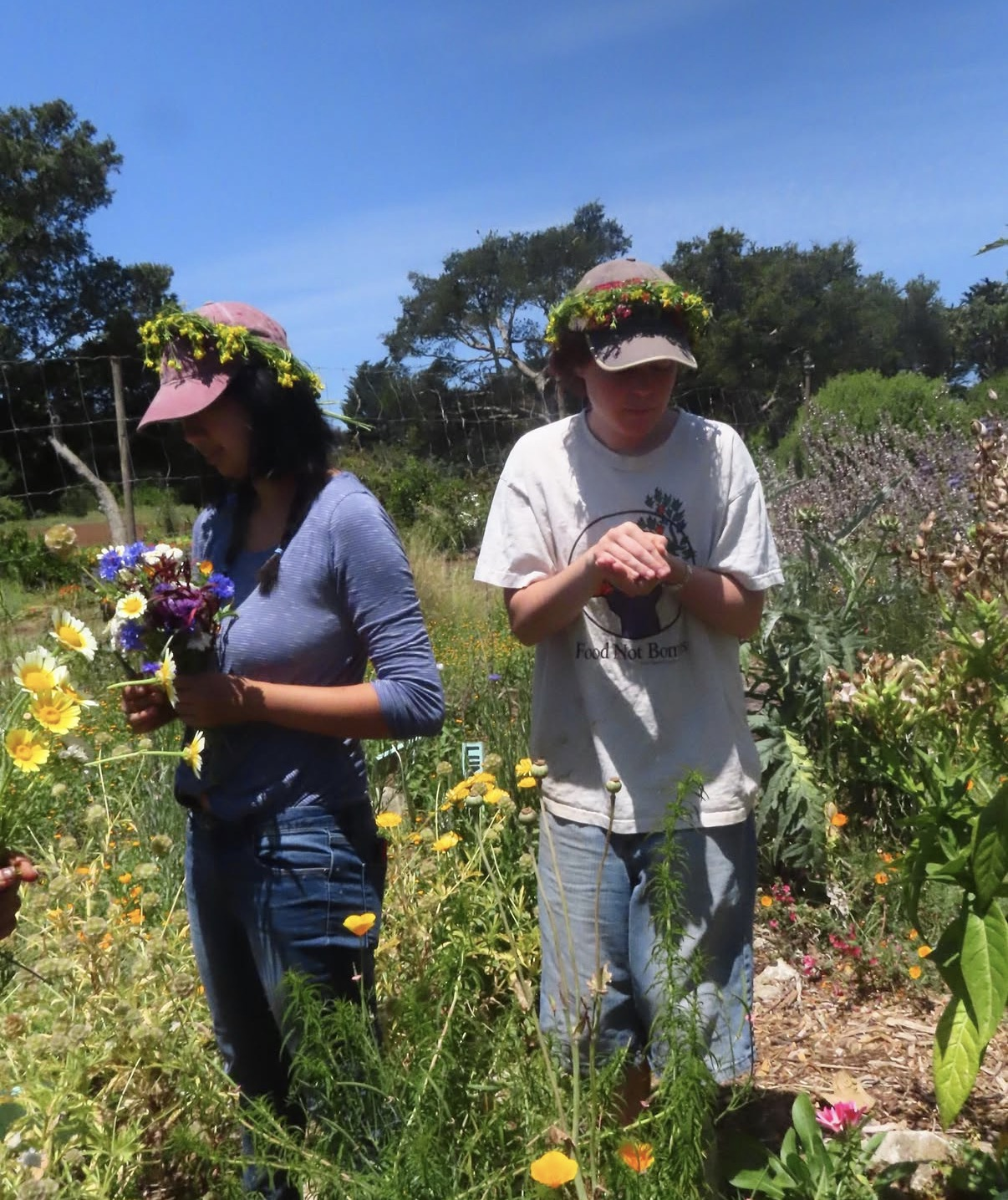 Two young women in a garden of colorful flowers, wearing hats decorated with flowers, one holding a bouquet and the other with hands clasped, during sunny weather.