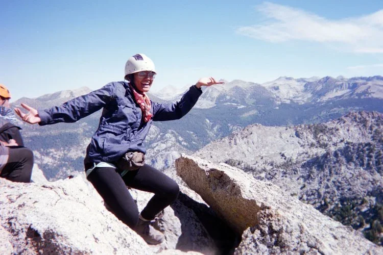 A woman wearing a helmet and sunglasses sitting on a rocky mountain peak with mountain ranges in the background.
