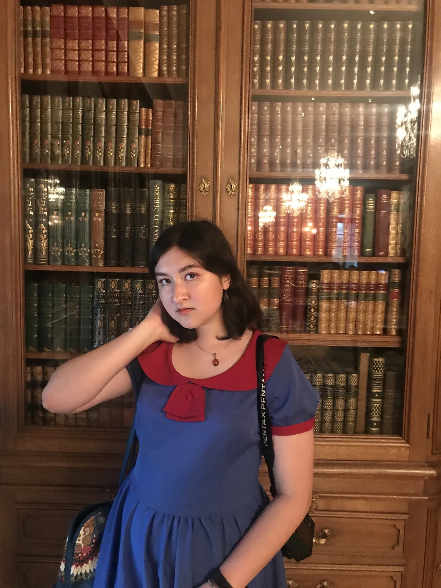 A young woman with shoulder-length dark hair, wearing a blue dress with red accents and a necklace, standing in front of a wooden bookshelf filled with old leather-bound books.