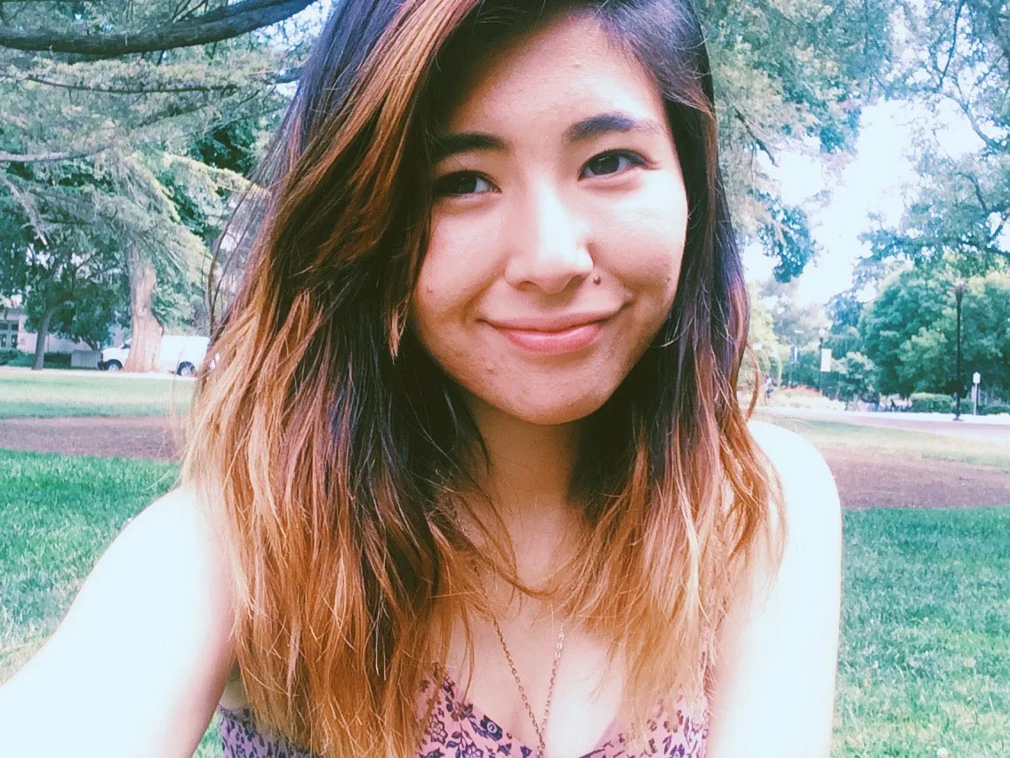 Smiling young woman with wavy, shoulder-length hair outdoors in a park with trees and grass.