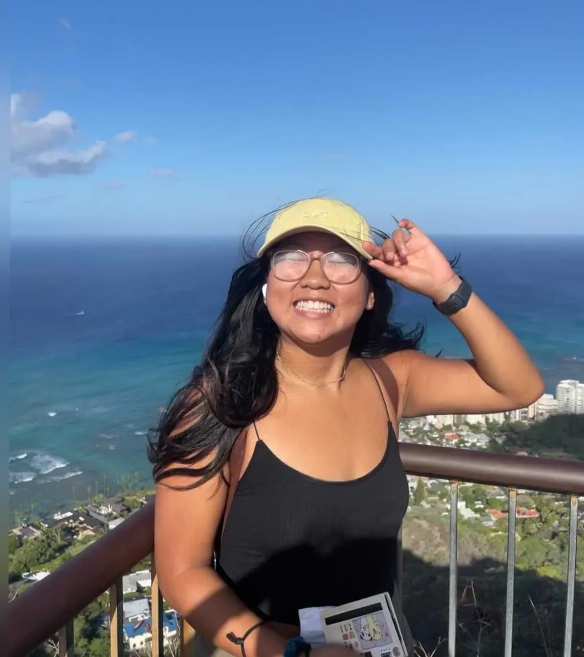 A woman smiling outdoors on a sunny day, wearing glasses, a yellow hat, a black tank top, and a watch, with ocean and cityscape in the background.