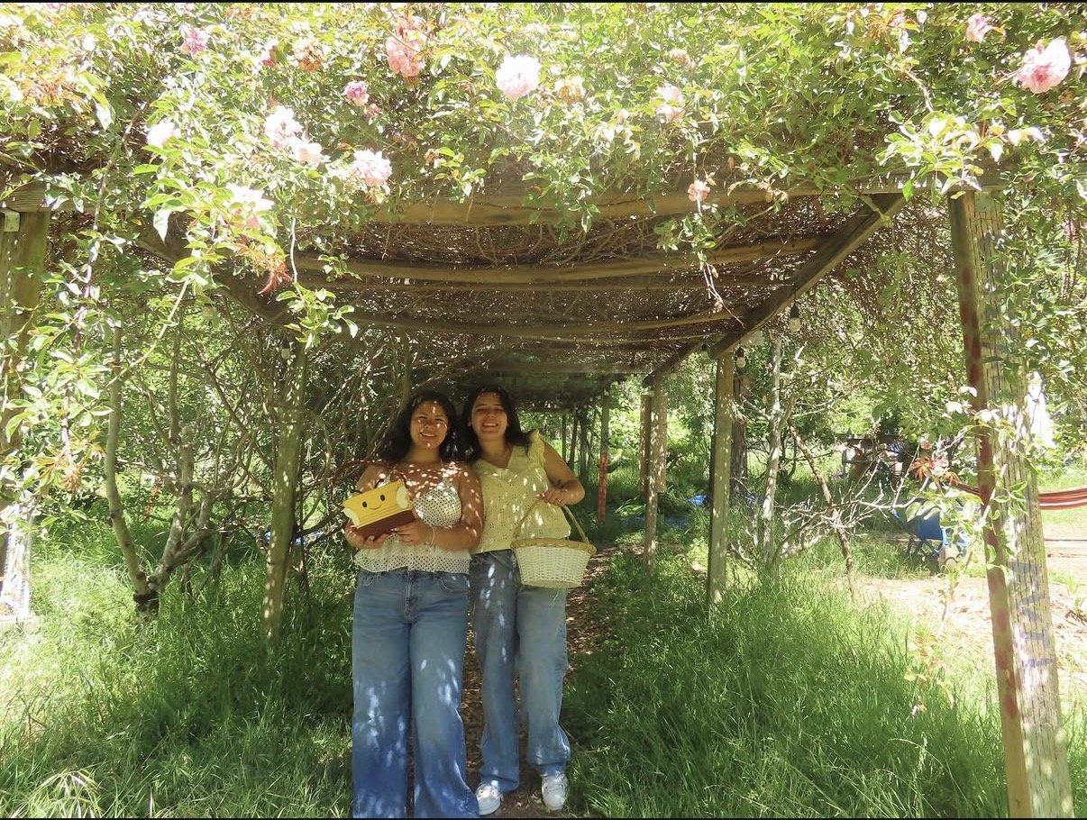 Two women smiling and posing for a photo under a wooden archway covered with pink and white flowers, holding a gift box and a basket, in a green garden setting.