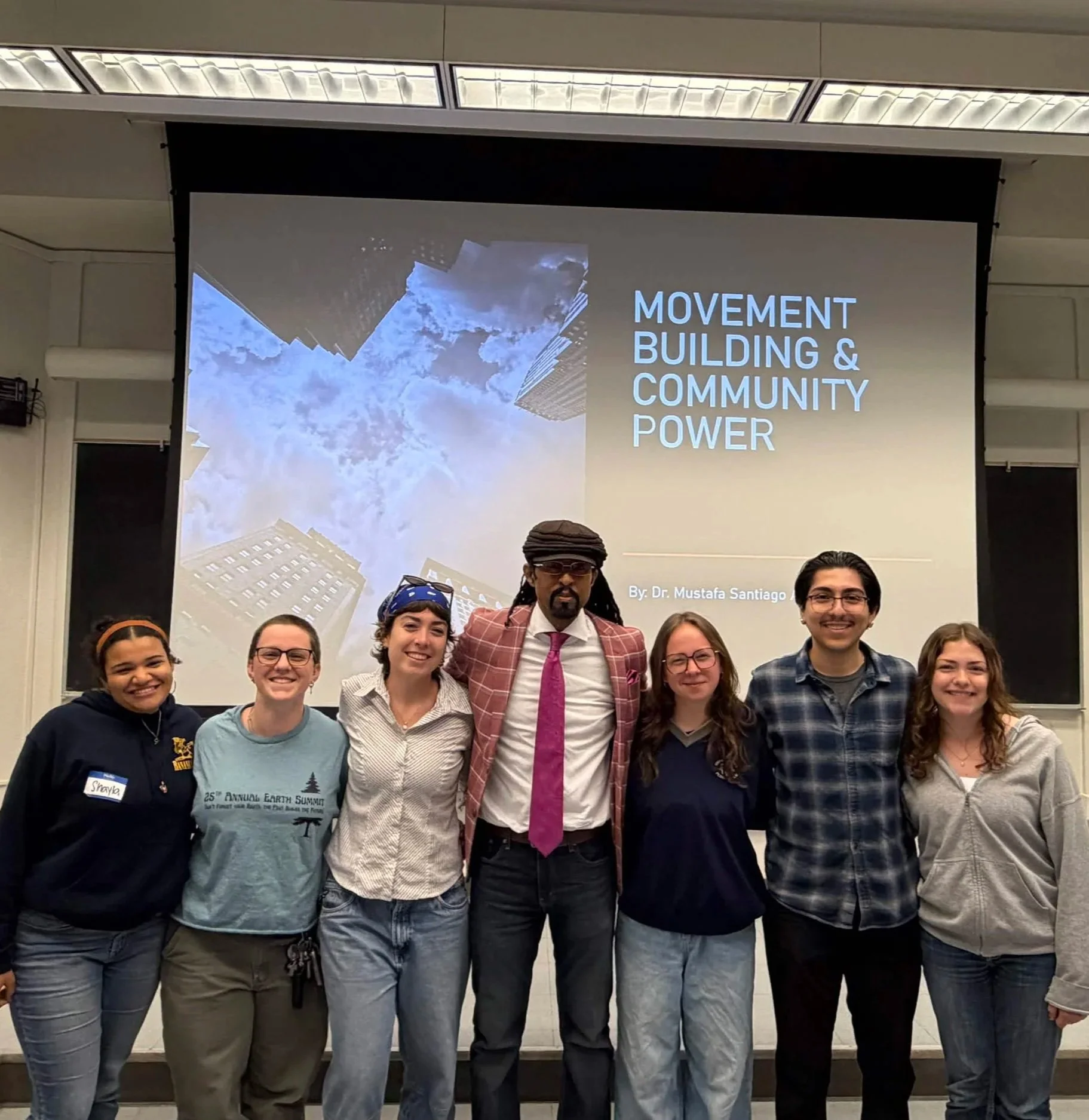 Group of people standing in front of a large screen displaying a presentation titled 'Movement Building & Community Power' at an event or conference.
