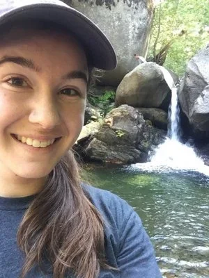 Smiling young woman taking a selfie outdoors near a waterfall and river with trees in the background.