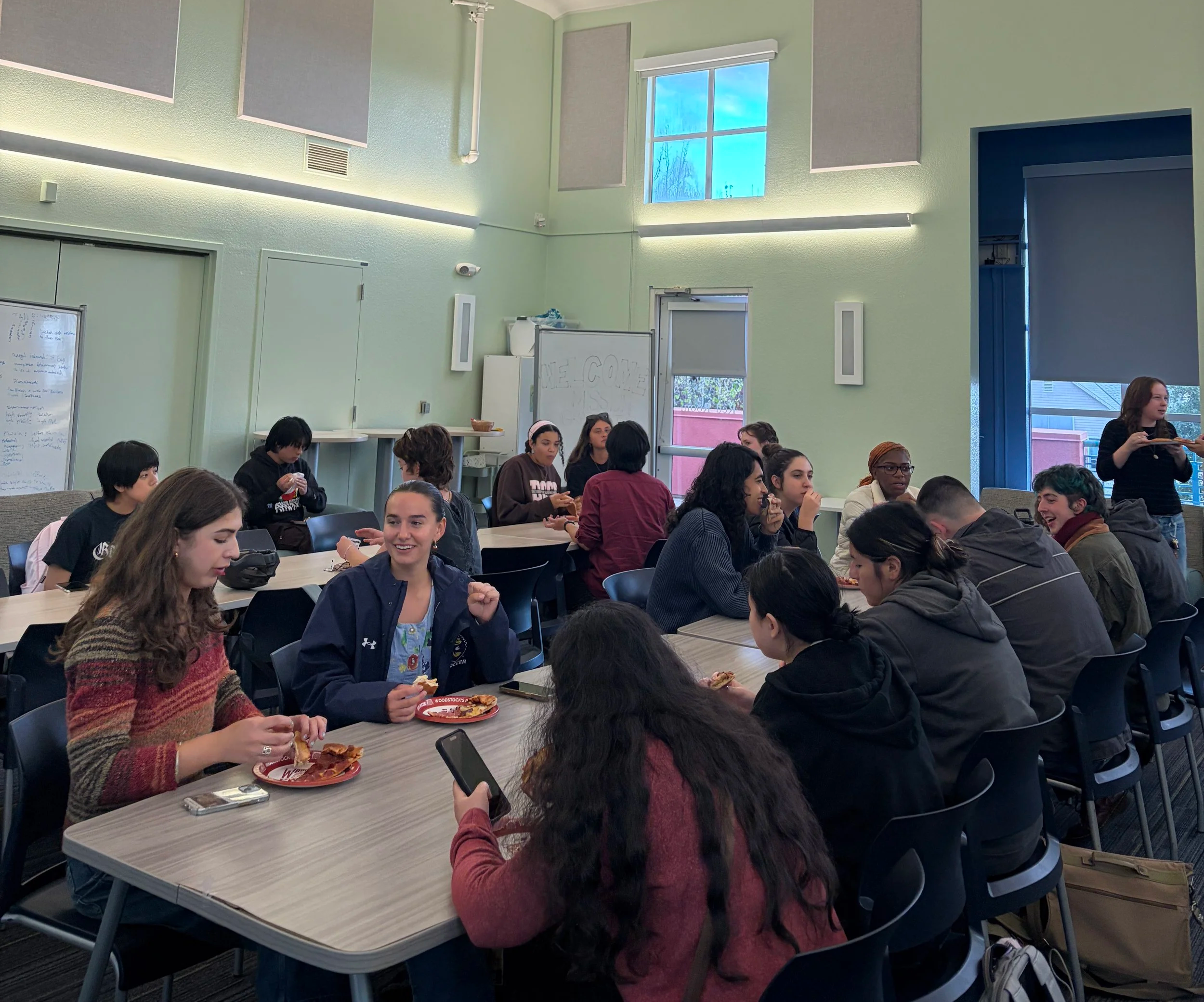 A group of diverse young people sitting around a large table in a brightly lit room with green walls, eating pizza and socializing. Some are engaged with their phones, while others are talking and smiling.