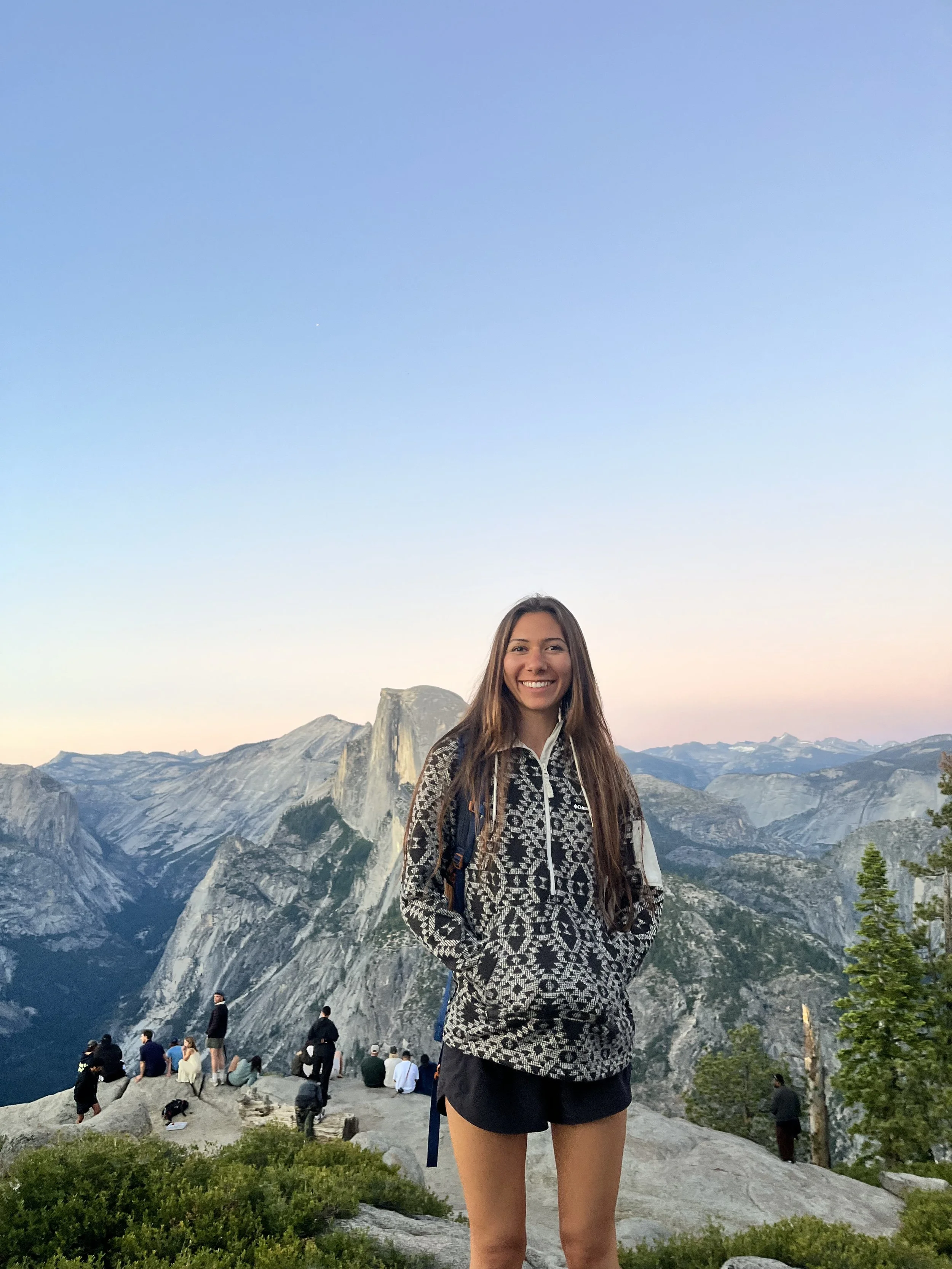 Young woman smiling at the Grand Canyon with arms outstretched, wearing a gray cap, red tank top, and denim shorts.