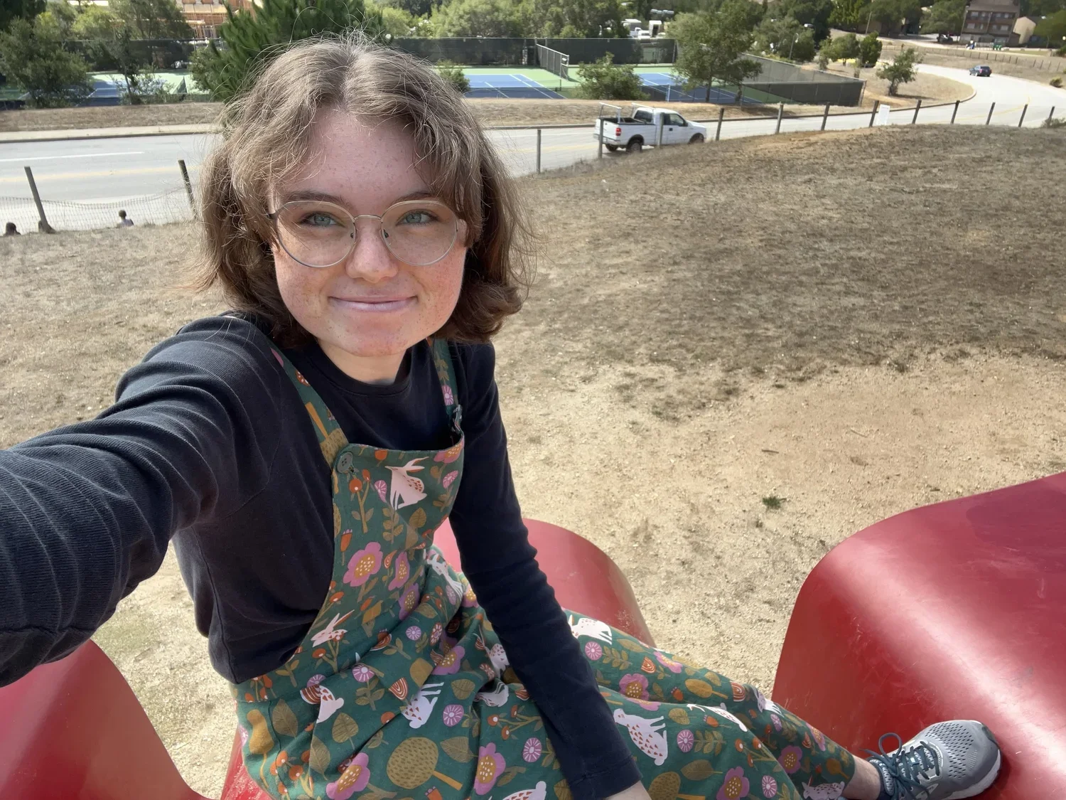 A young woman with glasses and shoulder-length wavy hair sitting on a red playground slide, smiling at the camera, in an outdoor park area with tennis courts, a road with a car, and trees in the background.