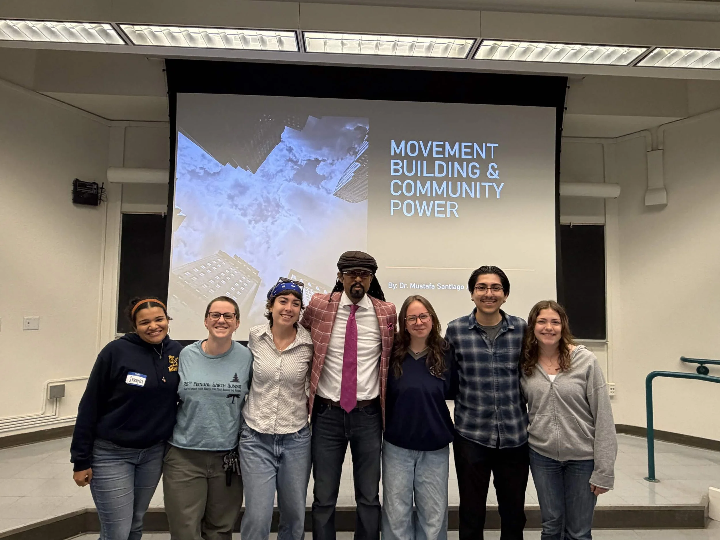 A group of seven people smiling and standing in front of a presentation screen that reads "Movement Building & Community Power." The group includes young adults and an older man with glasses, a hat, and a plaid blazer. The presentation appears to be at an indoor conference or seminar room.