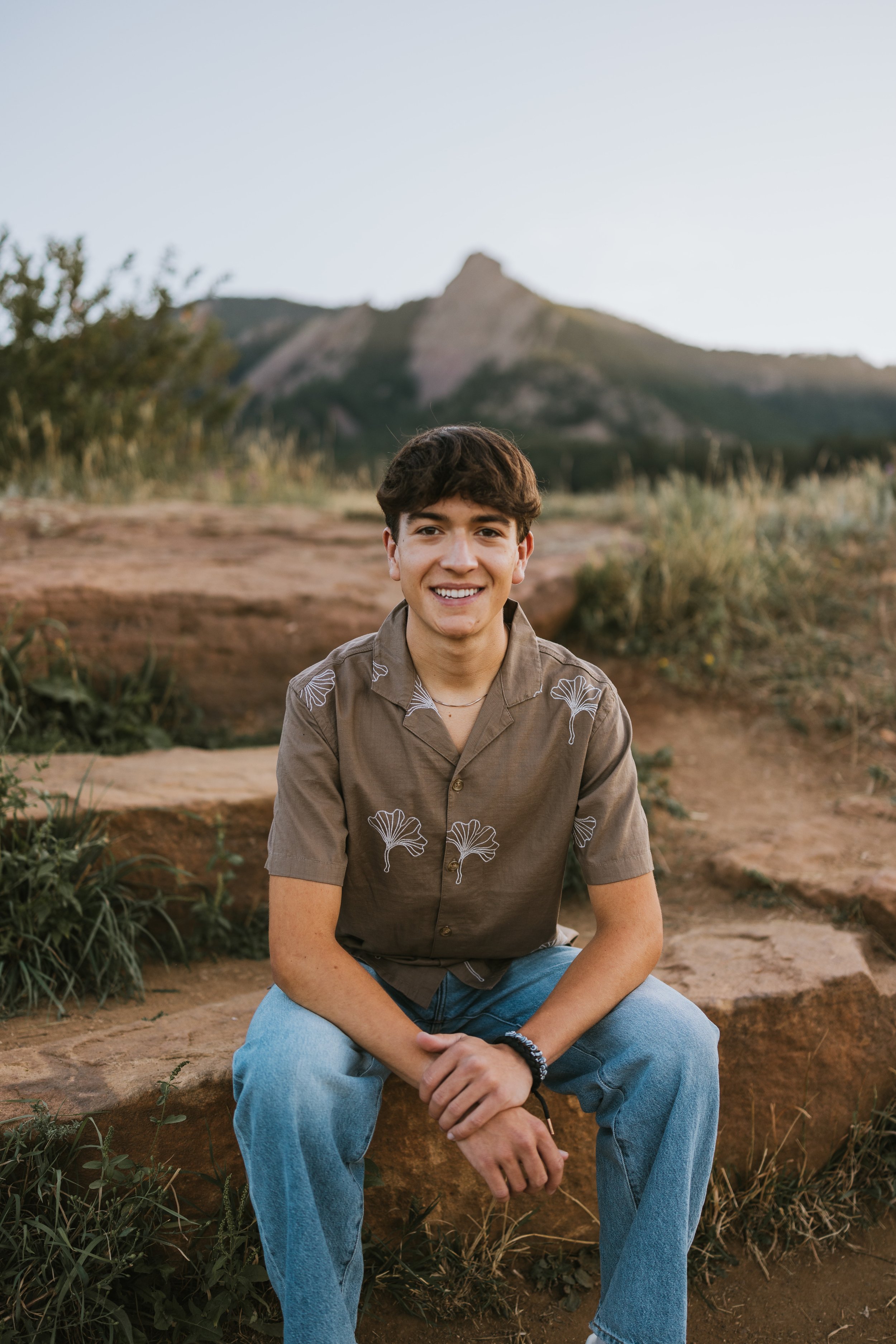 A young man sitting on rocks outdoors with mountains in the background, smiling, wearing a brown shirt with ginkgo leaves and blue jeans.