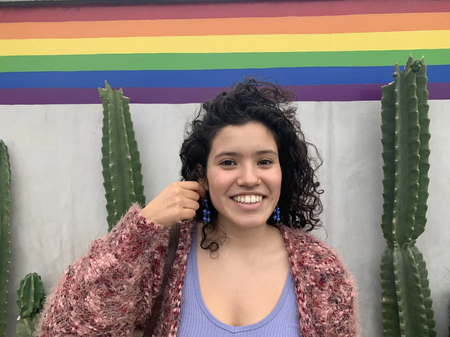 A smiling woman with curly dark hair, in a pink and purple textured sweater, wearing blue earrings, standing in front of a white wall with tall green cacti, and a rainbow painted on the wall above.
