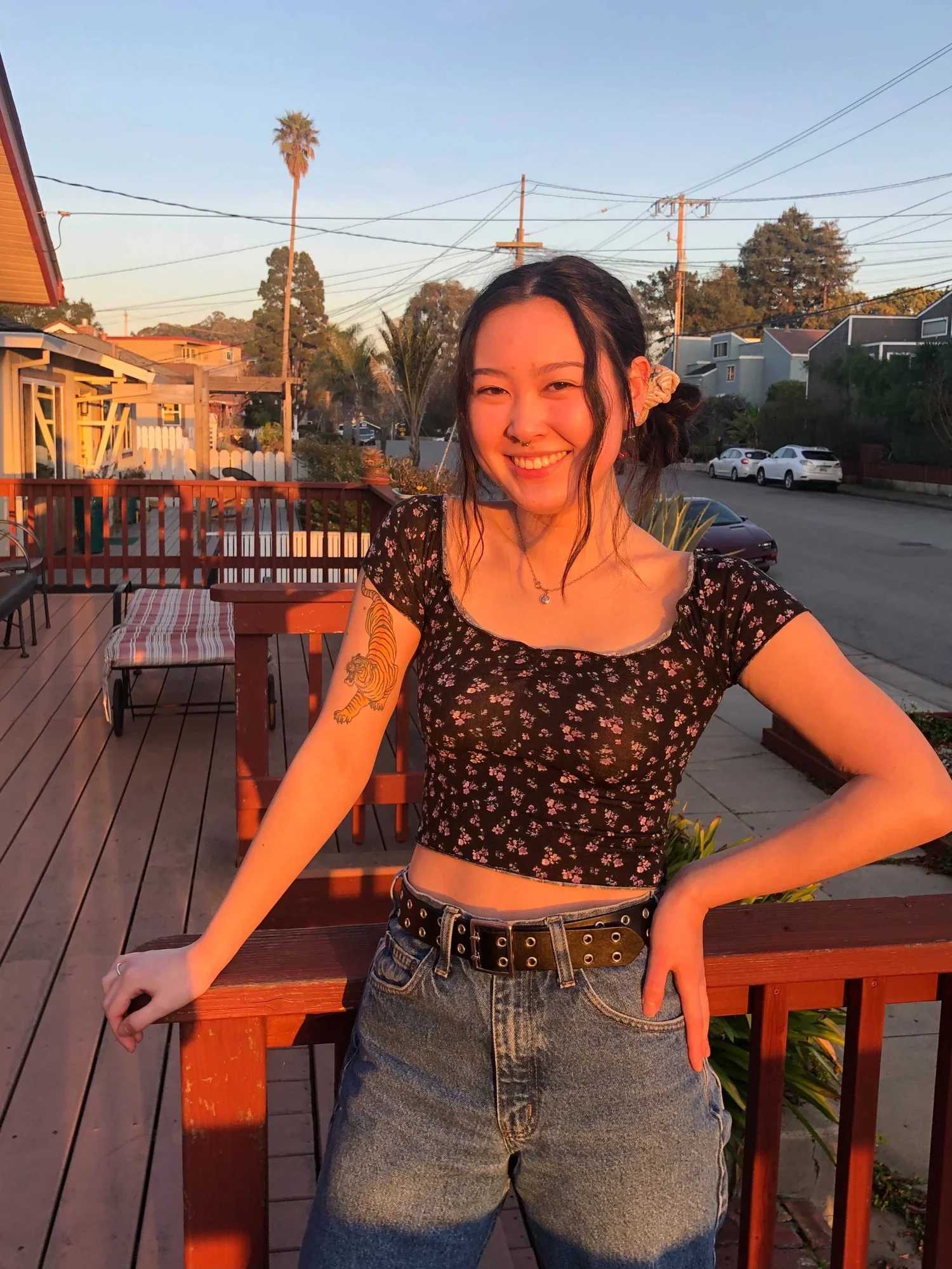 A young woman with dark hair, smiling, wearing a black floral crop top and blue jeans, standing outdoors on a wooden deck during sunset, with trees, houses, and parked cars in the background.