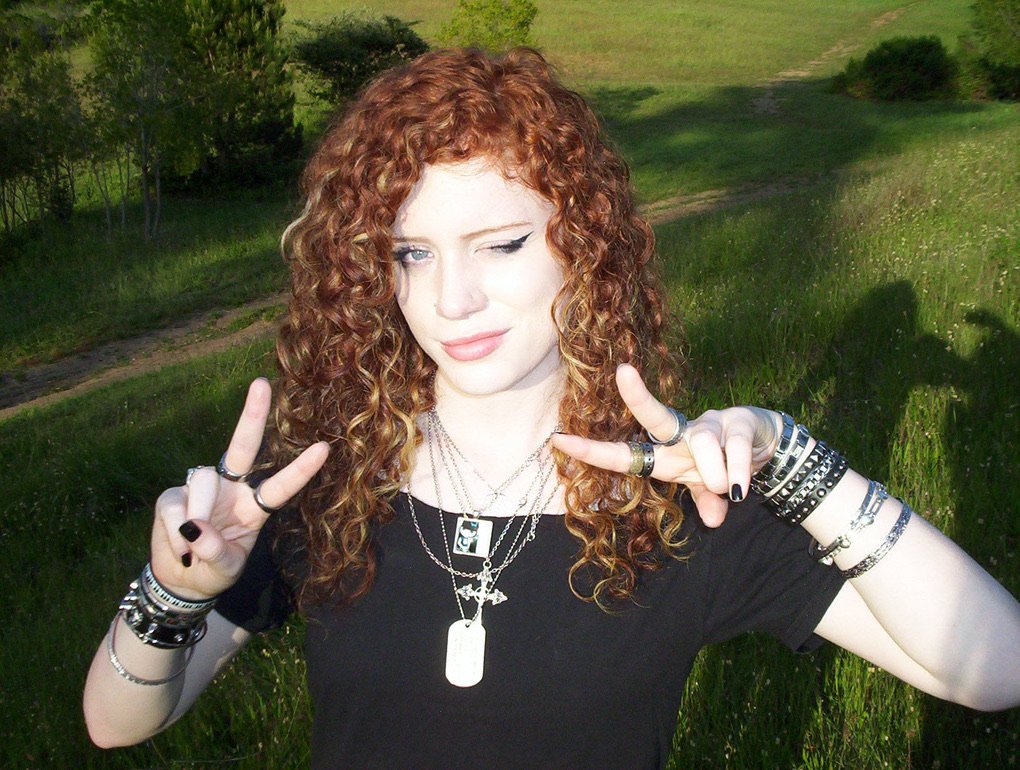 Young woman with curly red hair taking a selfie outdoors in a green grassy area, making peace signs with both hands.