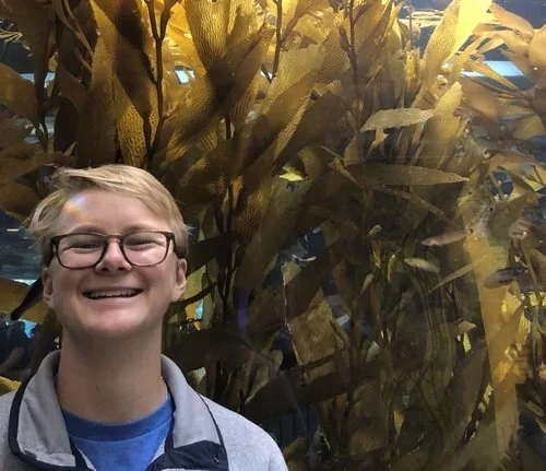 A smiling young person with glasses and blonde hair standing in front of large, tall plants or foliage.