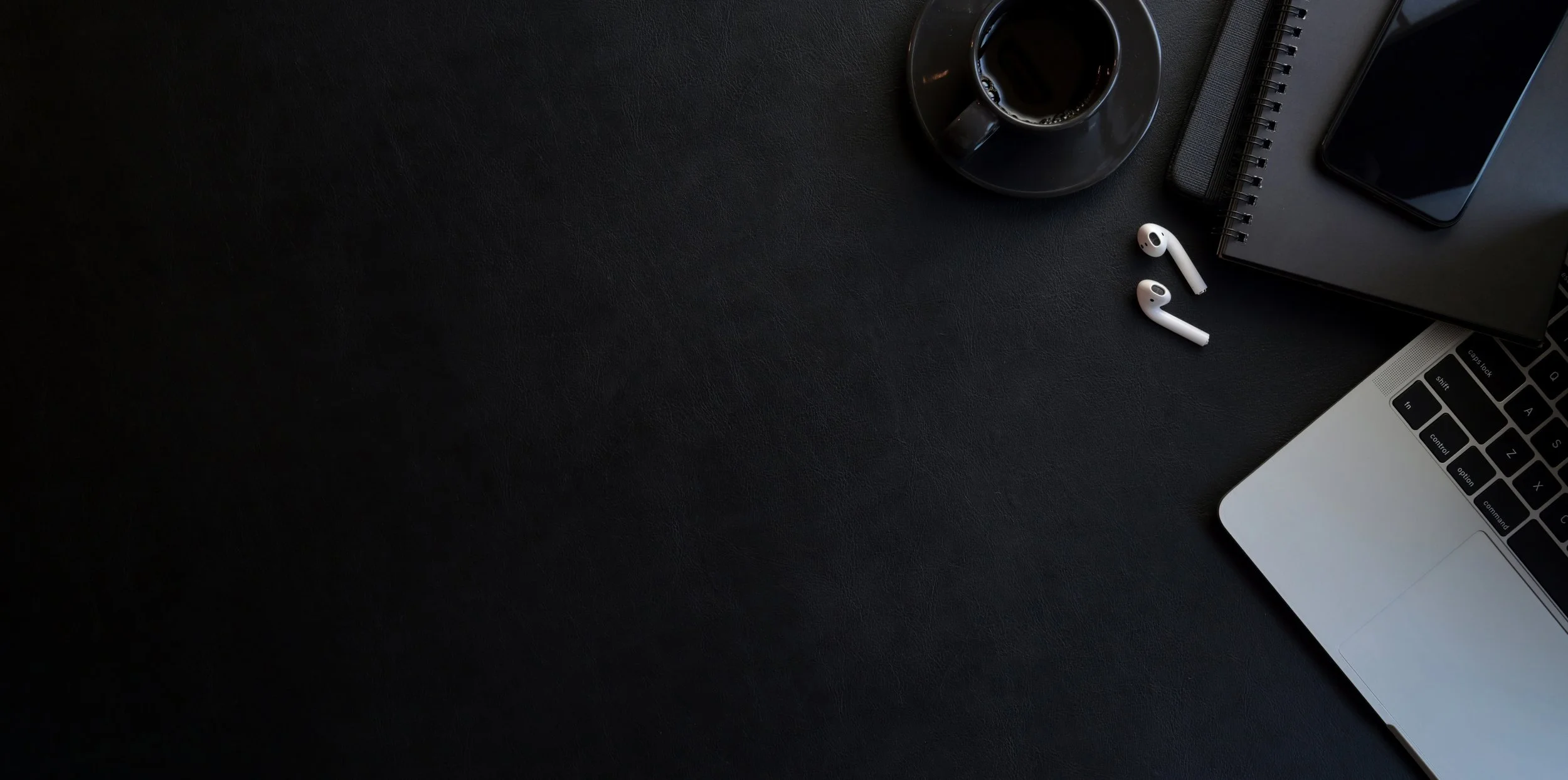 Top view of a black desk with a cup of black coffee, wireless earbuds, a closed notebook, a smartphone, and a laptop with the keyboard visible.