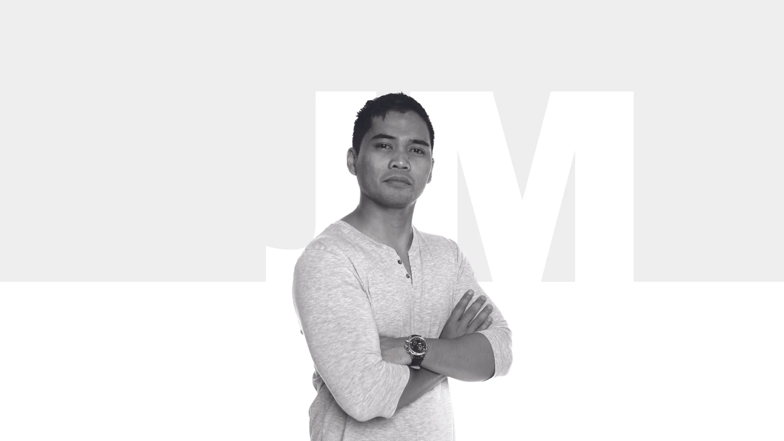 Black and white photo of a man with crossed arms, wearing a light long-sleeve shirt and a watch, standing in front of a background with large partially visible letters.