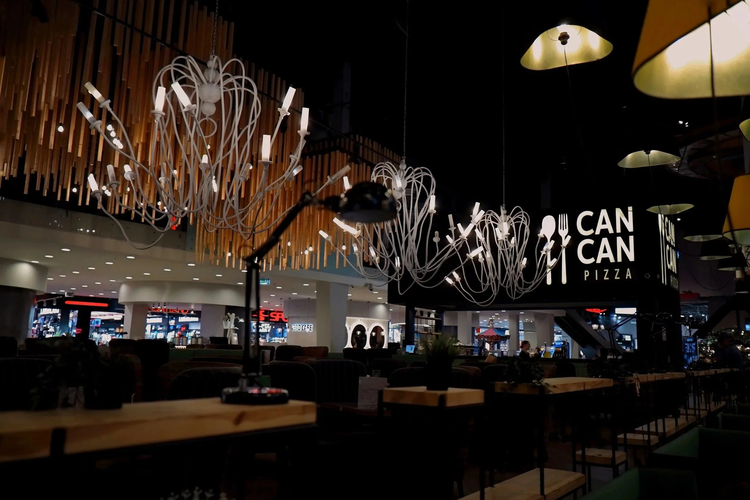 Innenansicht eines Restaurants, erkennbar an den Lampen und dem großen Schild mit »CAN CAN PIZZA«. Es sind Tische und Stühle zu sehen, sowie eine dunkle Decke und dekorative Luster.