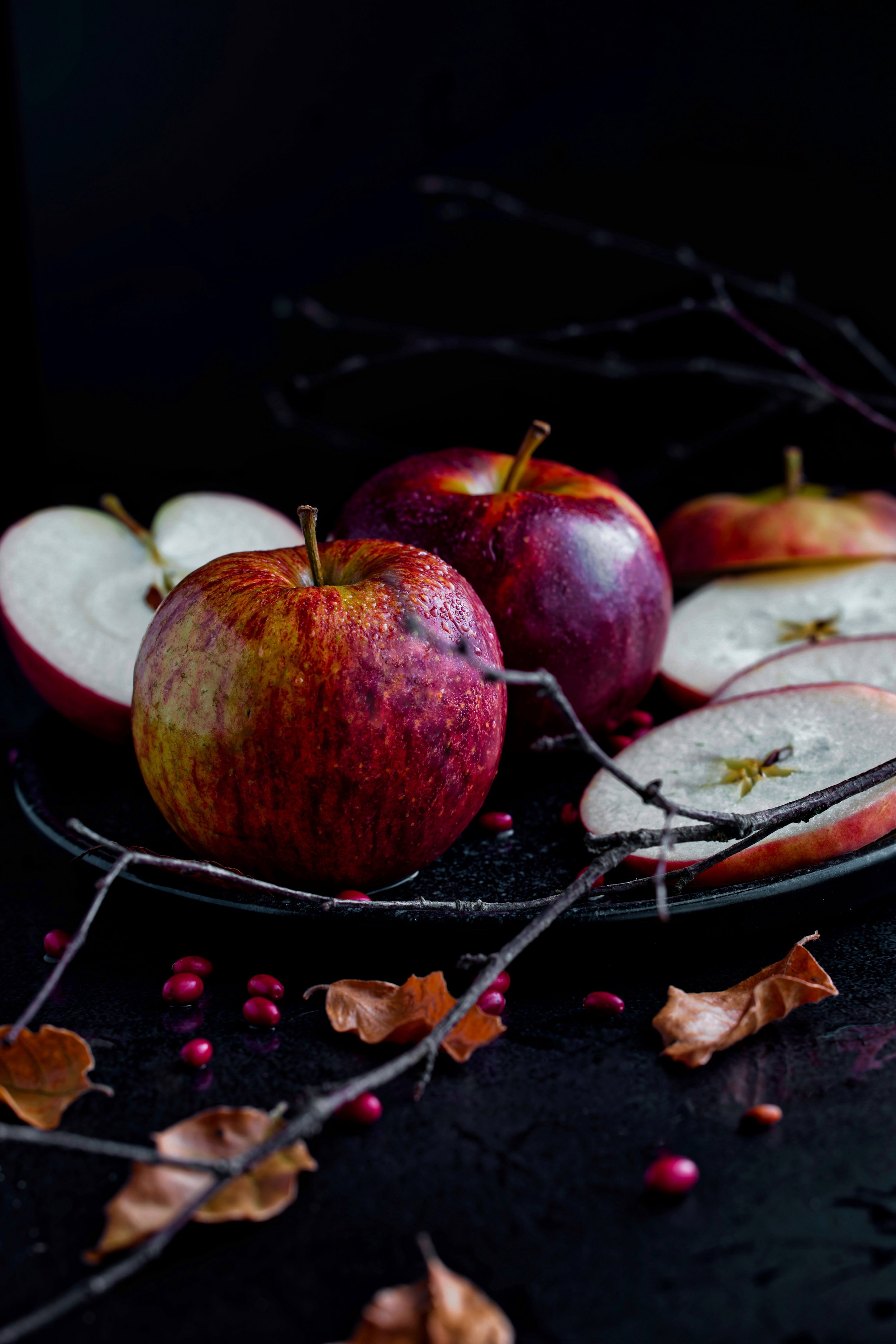 Drei rote Äpfel, eine Hälfte eines Apfels und einige trockene Blätter und Zweige auf einem dunklen Teller und Hintergrund.