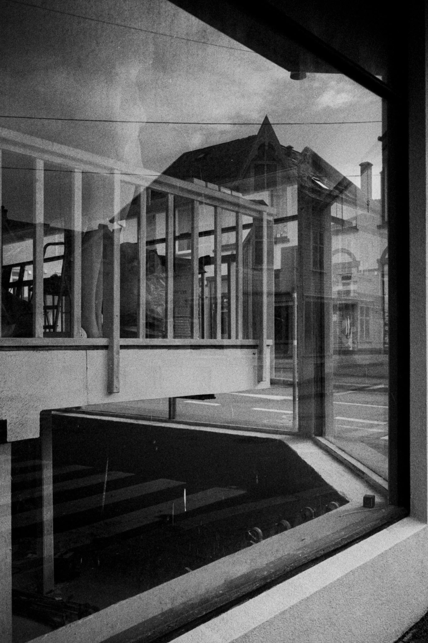 A black and white photo of a view through a window showing a construction site with unfinished or framed building structures, including wooden beams and a roof, and neighboring buildings in the background.