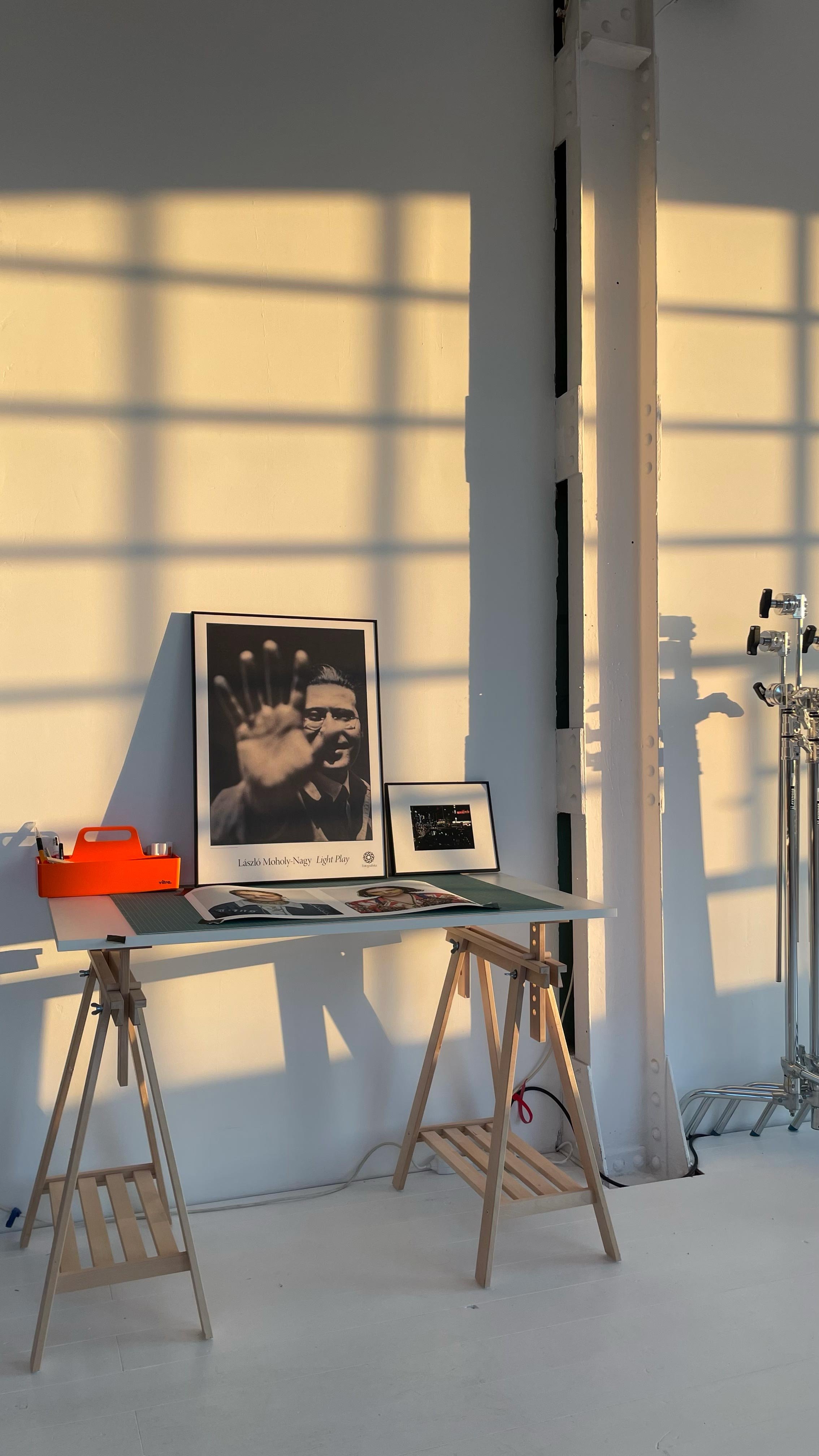 A desk with framed photographs and magazines, illuminated by sunlight with shadows on the wall, beside studio equipment.