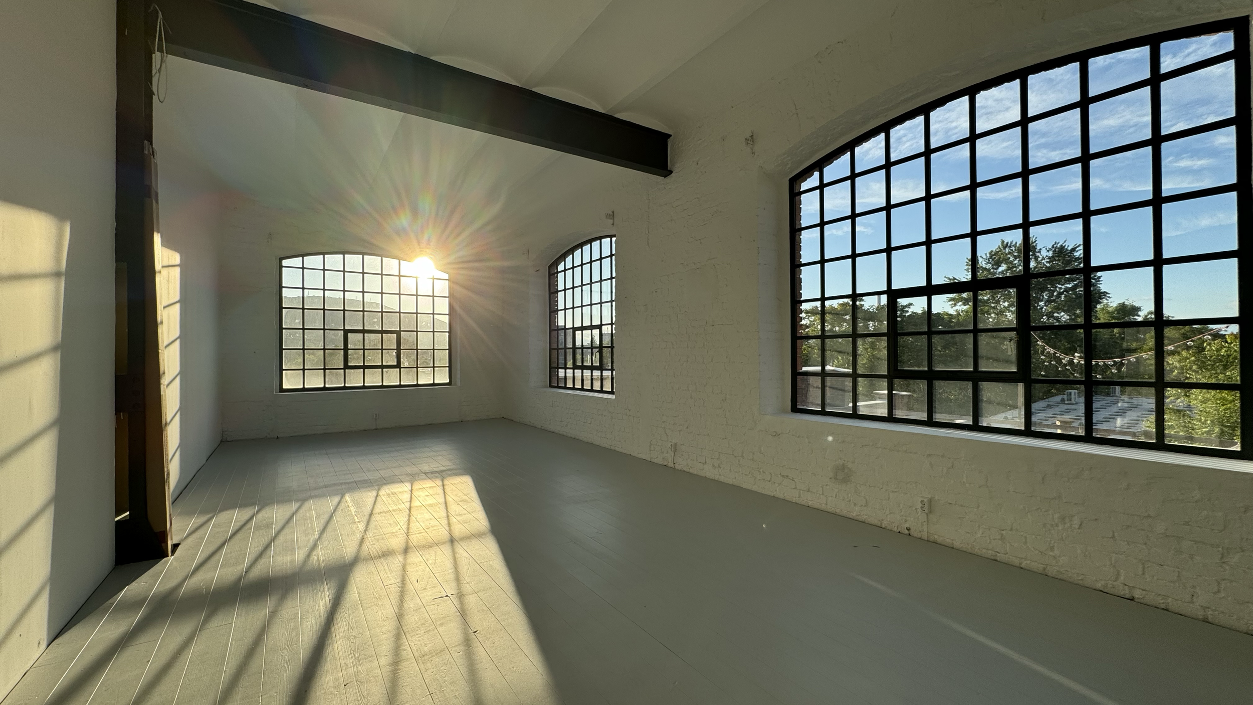 Empty room with large industrial-style windows allowing sunlight to stream in, casting shadows on light-colored wooden floors and white brick walls.