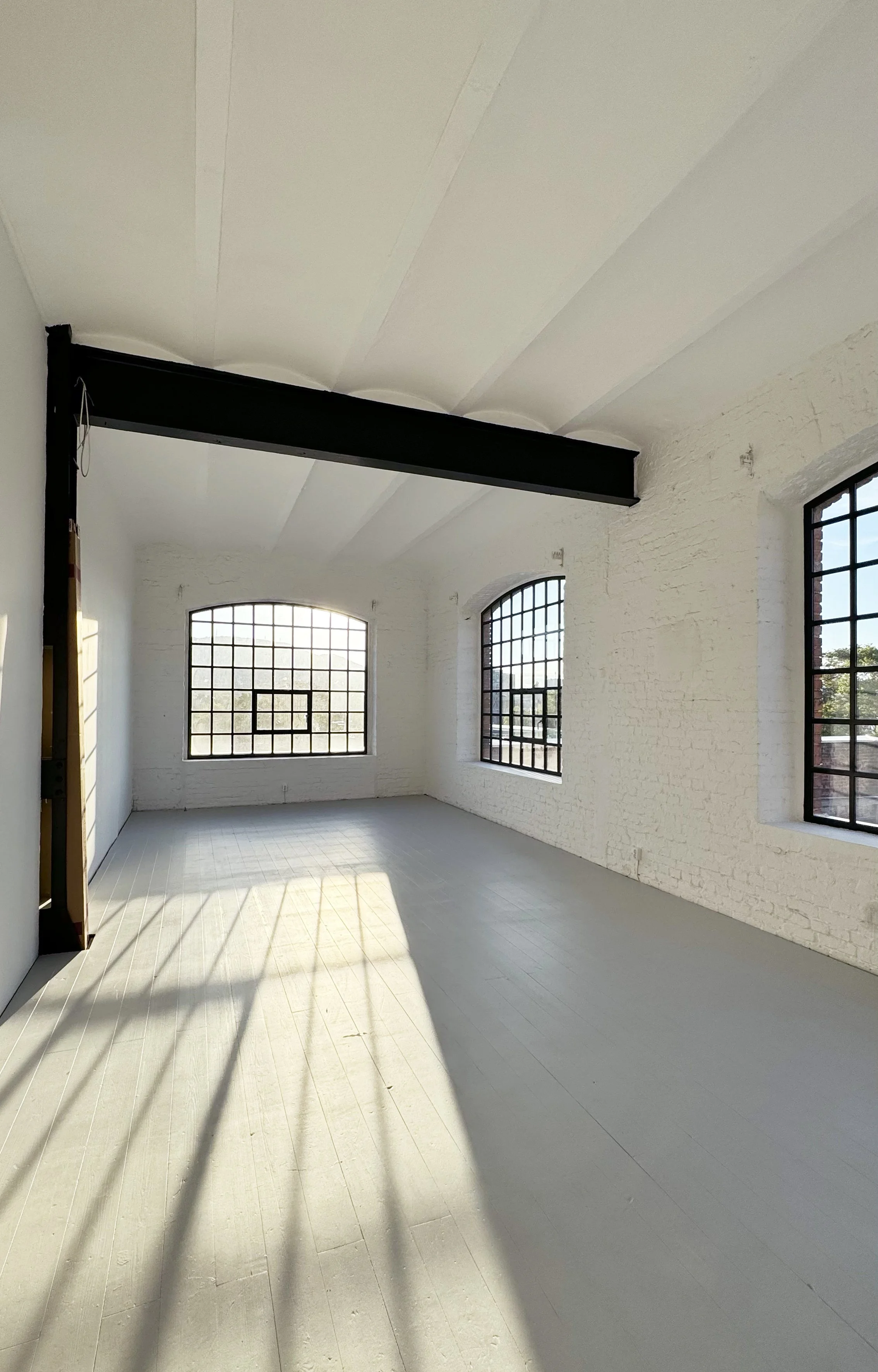 Empty loft-style room with white brick walls, large grid windows, gray painted wooden floors, and a black metal beam across the ceiling, with sunlight casting shadows on the floor.