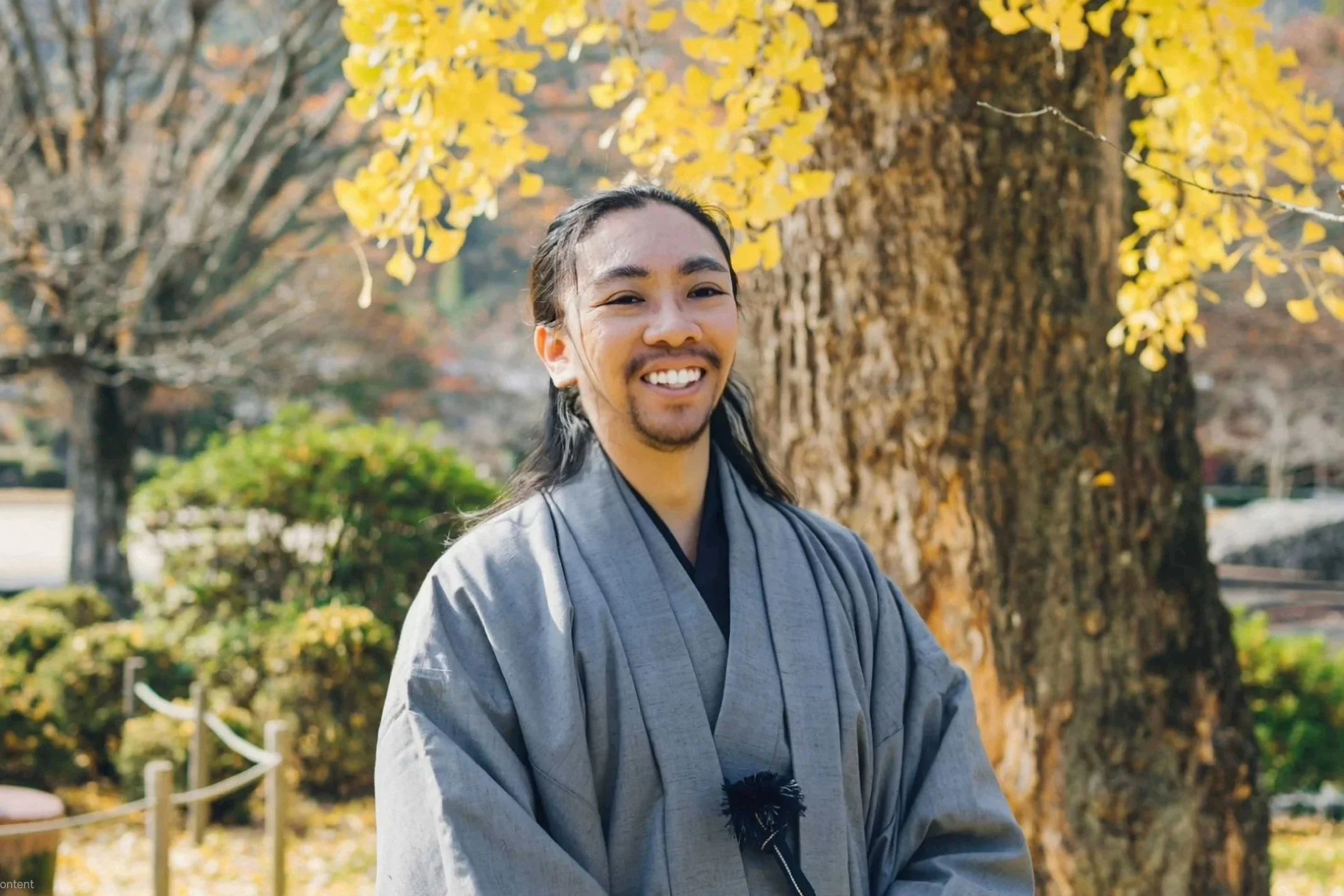 A man with long dark hair, a mustache, and beard smiling outdoors in front of a large tree with yellow leaves, wearing traditional gray clothing.