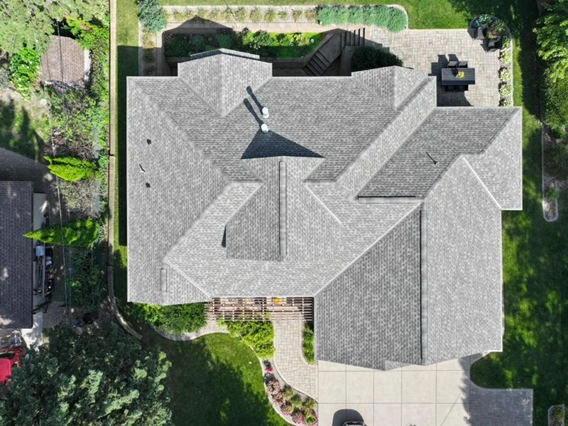 An aerial view of a house with multiple gabled roofs, a driveway, green lawn, and surrounding trees.