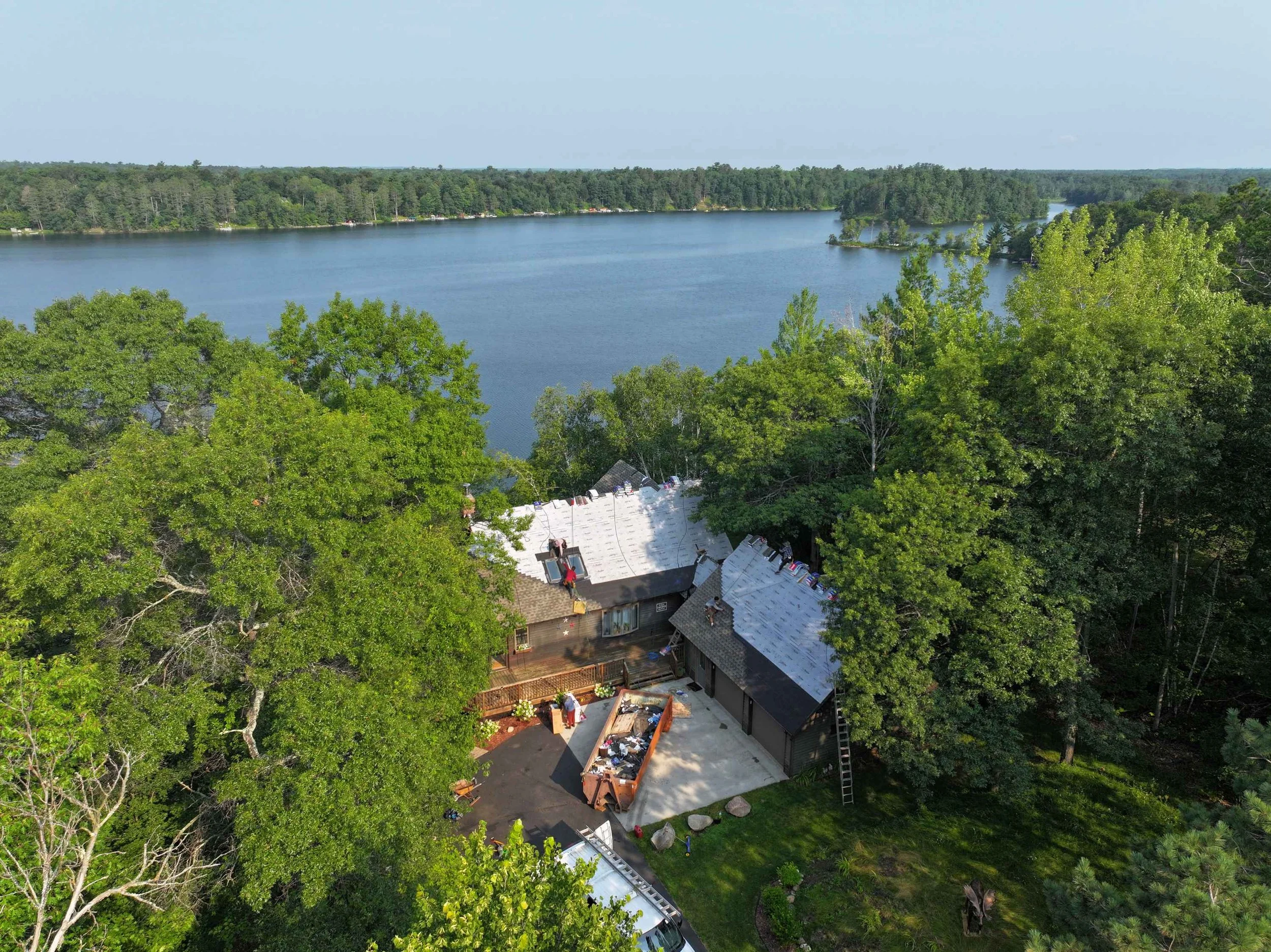 Aerial view of a lake surrounded by dense green trees and a house with a white roof in the foreground.