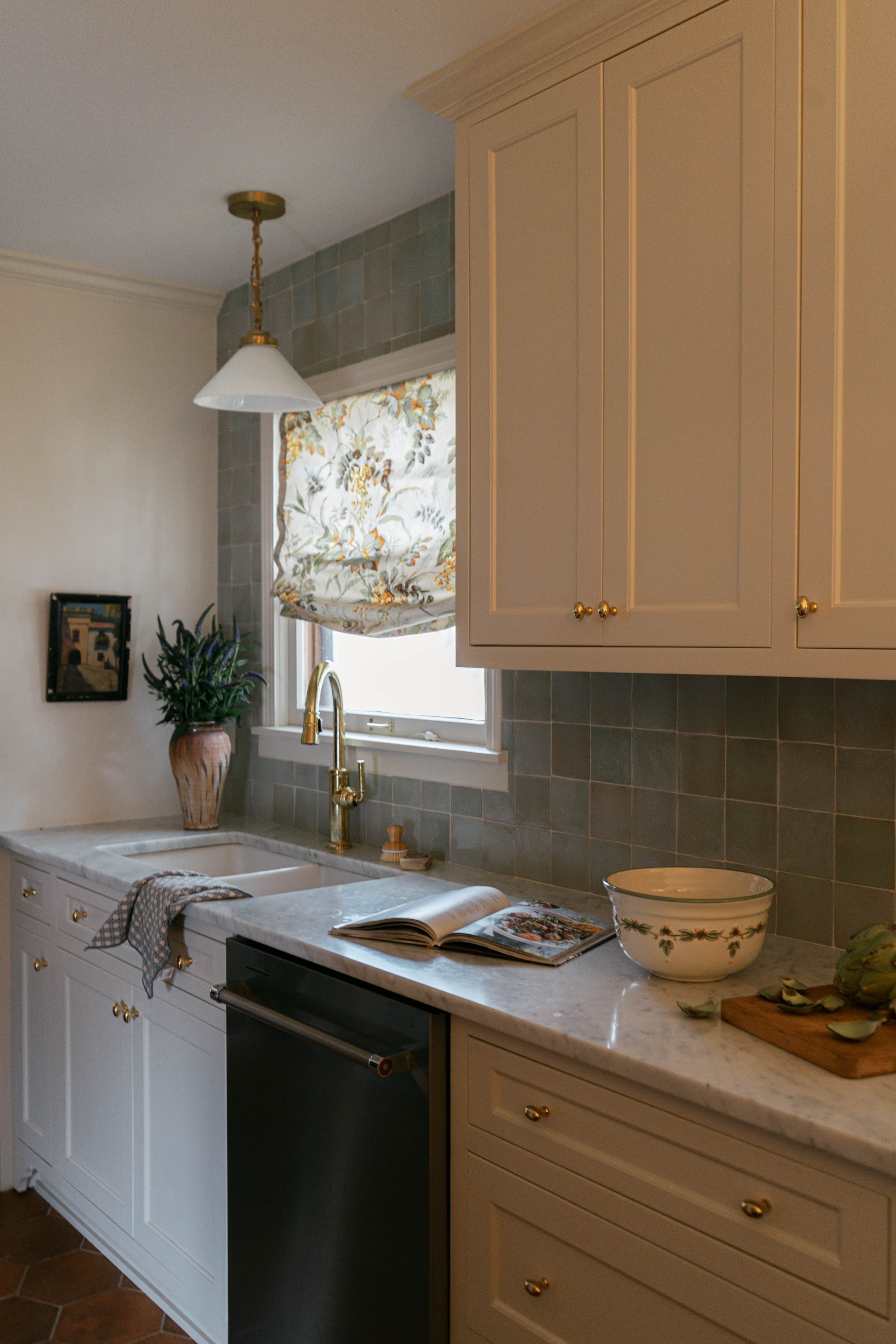 A cozy kitchen corner with beige cabinets, a white marble countertop, a large bowl with a floral pattern, an open cookbook, a cutting board with avocado slices, and a window with a floral valance. There is a gold faucet and a potted plant nearby.
