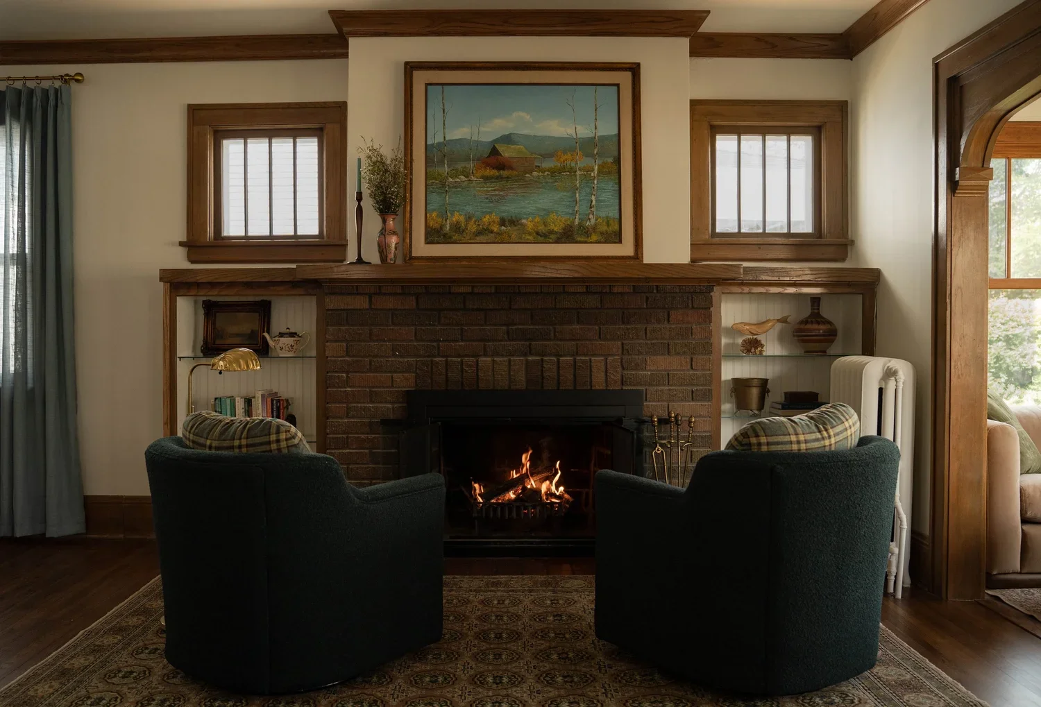 Living room with a brick fireplace, two plaid armchairs facing the fire, and a painting of a landscape above the mantle.