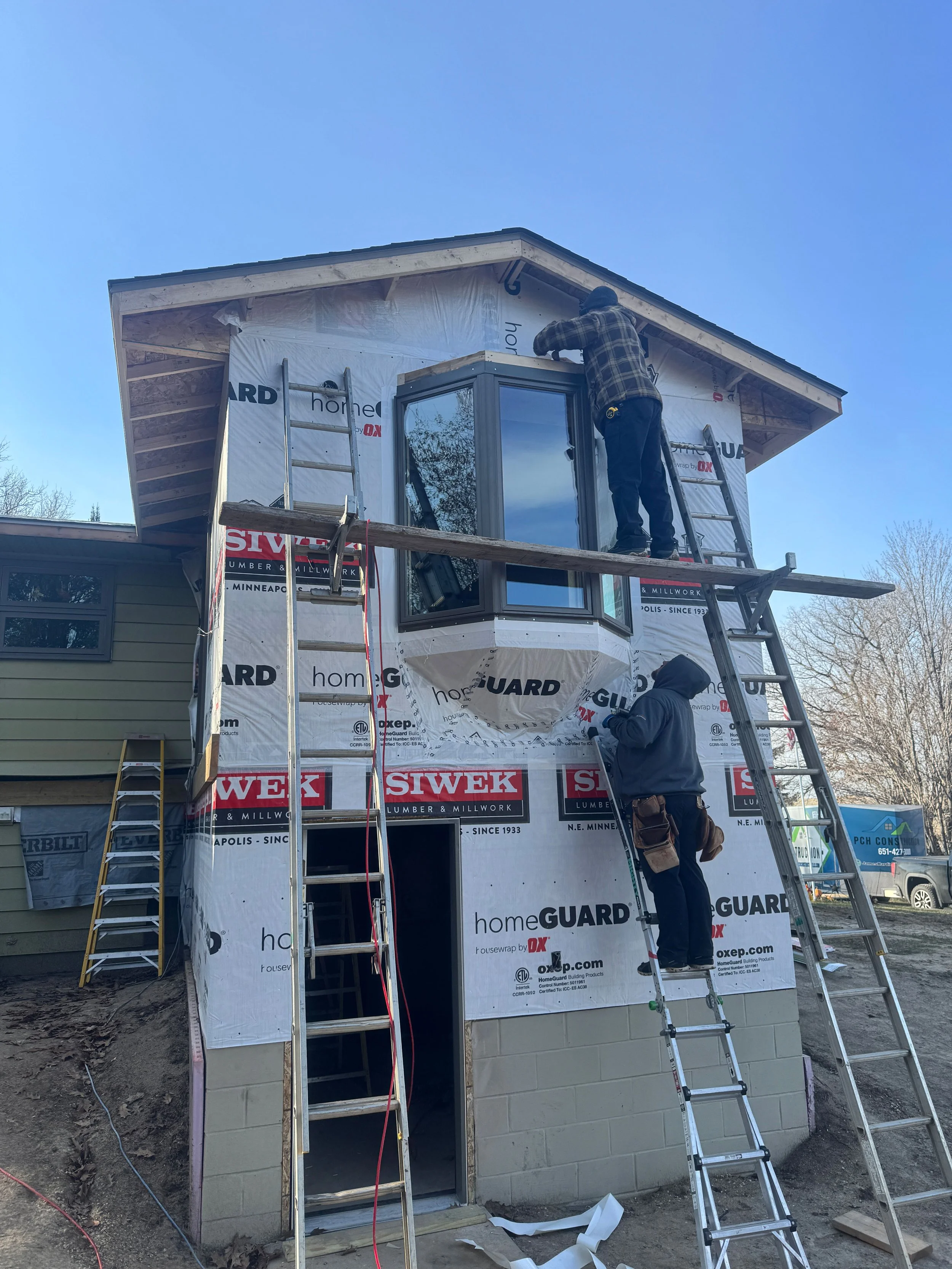 Construction workers installing or repairing a small building or extension on a house, with ladders and building materials visible.