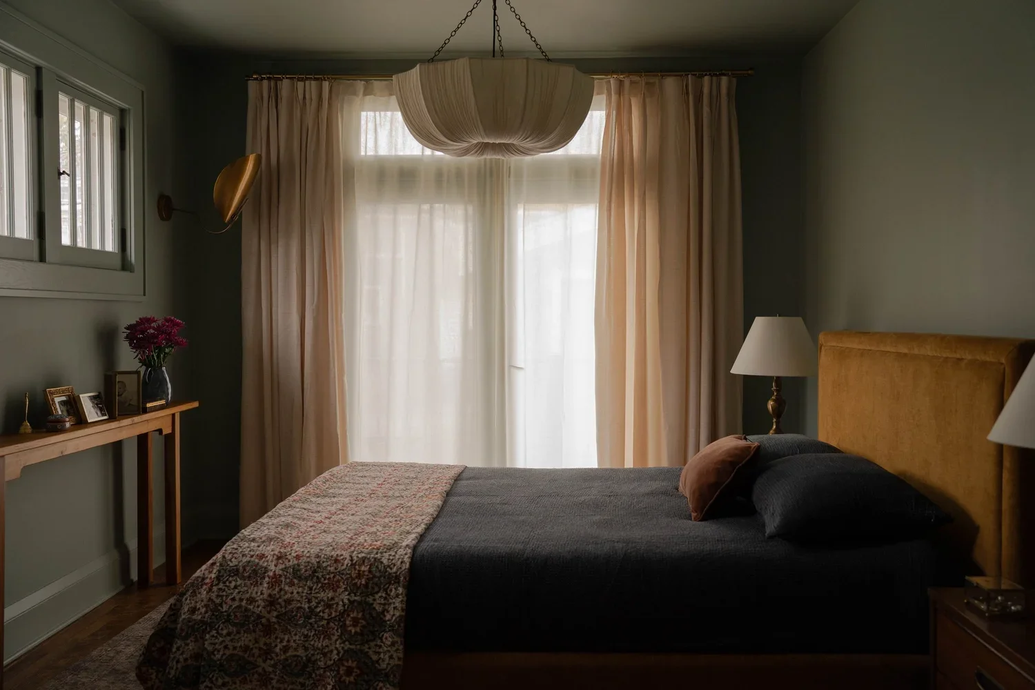 A cozy bedroom with a large window covered by sheer white curtains and beige drapes, a bed with dark bedding and beige headboard, two bedside lamps, a wooden nightstand, and a wooden console with framed photos and a vase of pink flowers.